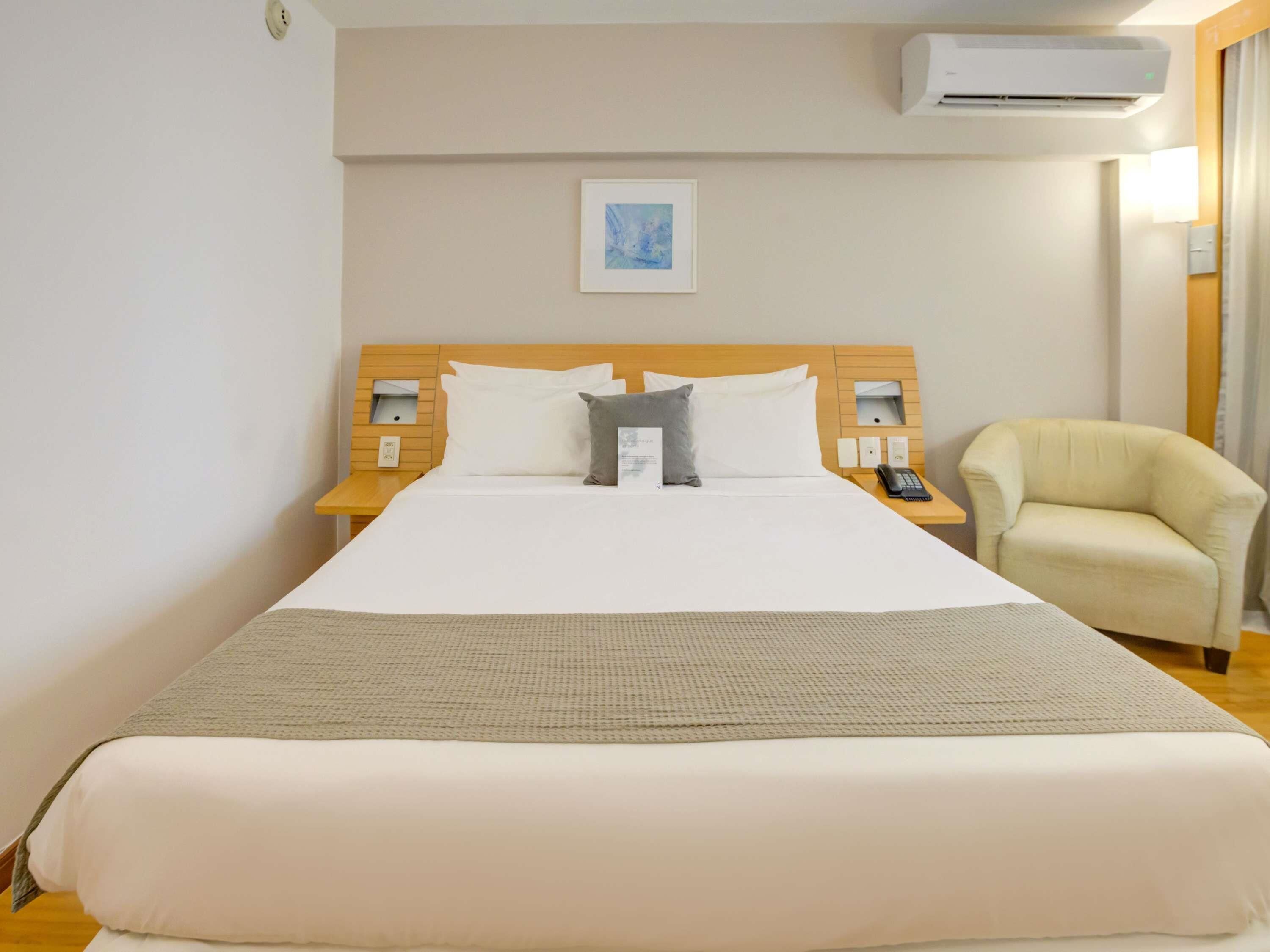 A hotel room with a large bed, a bedside table with a lamp, a chair, and an air conditioning unit on the wall.