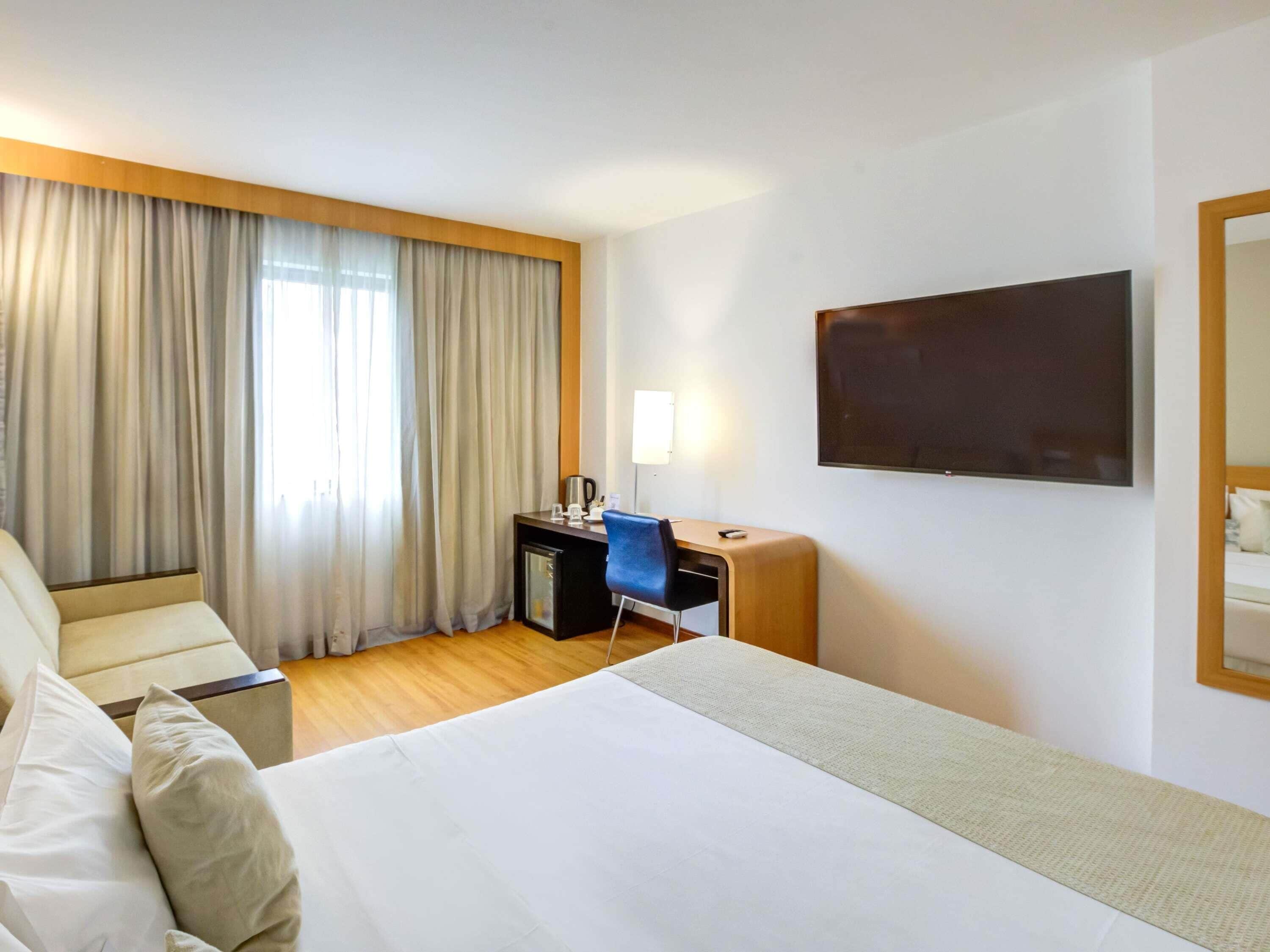 A hotel room with a bed, a desk, a chair, a TV, and curtains.