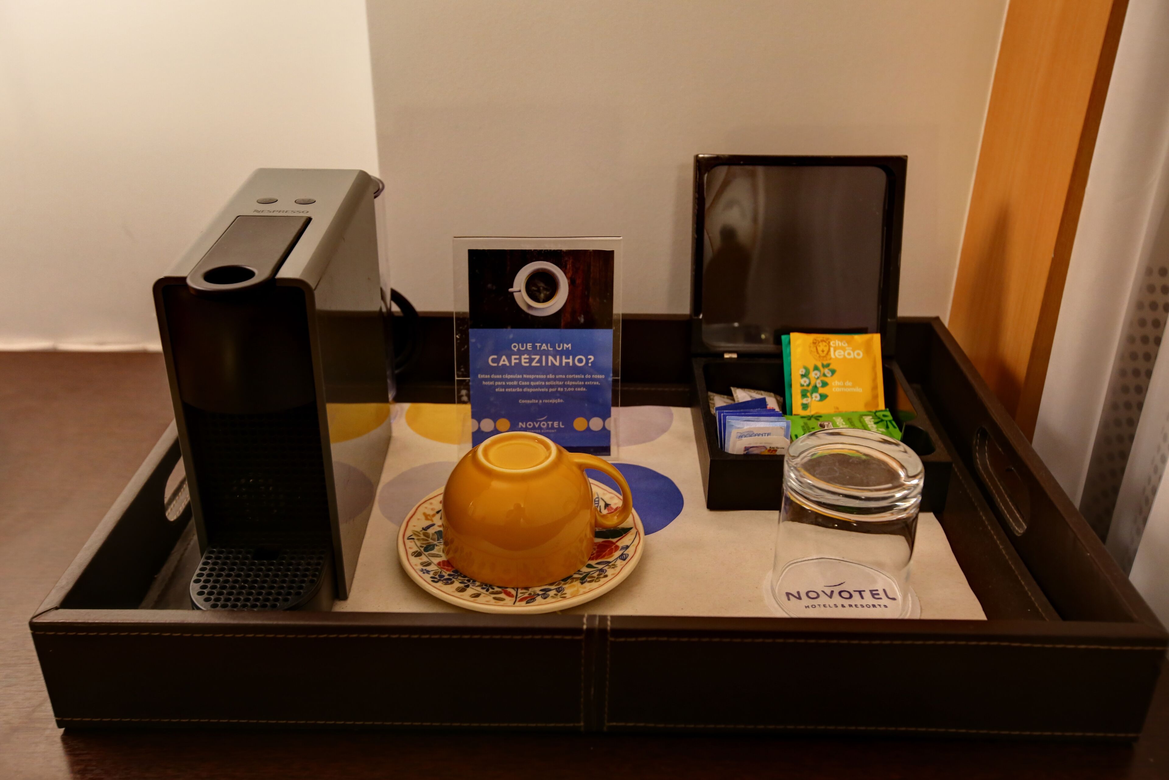 A minibar setup with a coffee machine, a cup of coffee, a coffee pod, a jar, and a Novotel branded item.