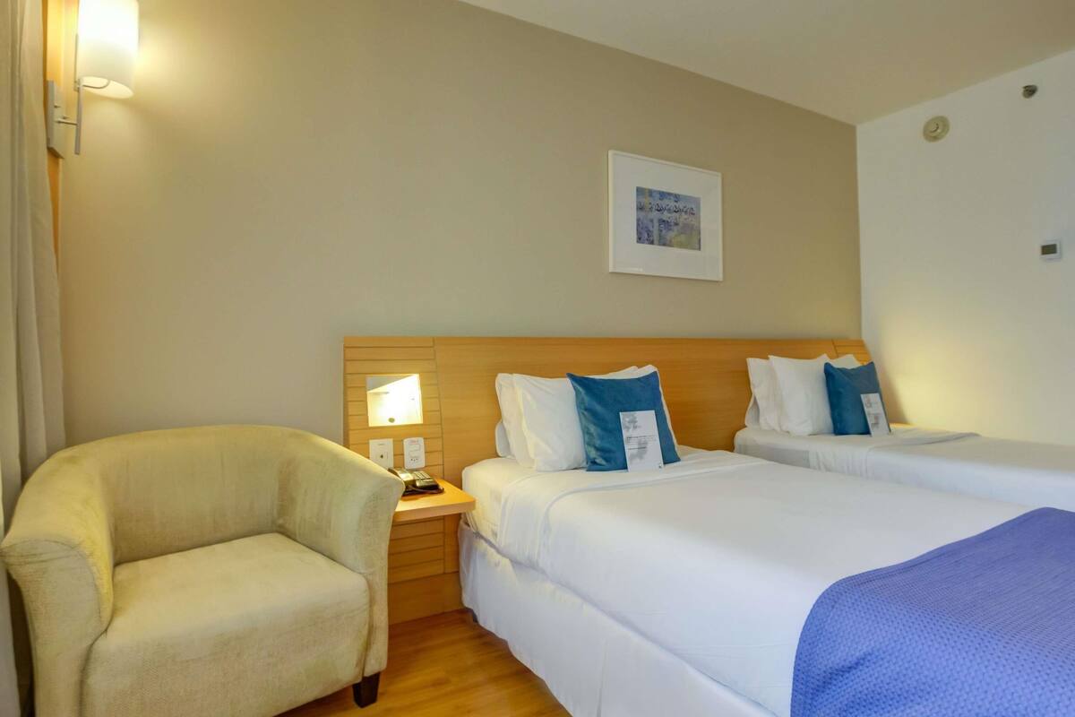 Habitación de hotel con cama, mesita de noche, lámpara, silla y un cuadro en la pared.