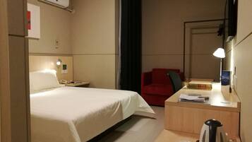 Habitación de hotel con cama, escritorio con lámpara, una silla roja, cafetera y espejo.