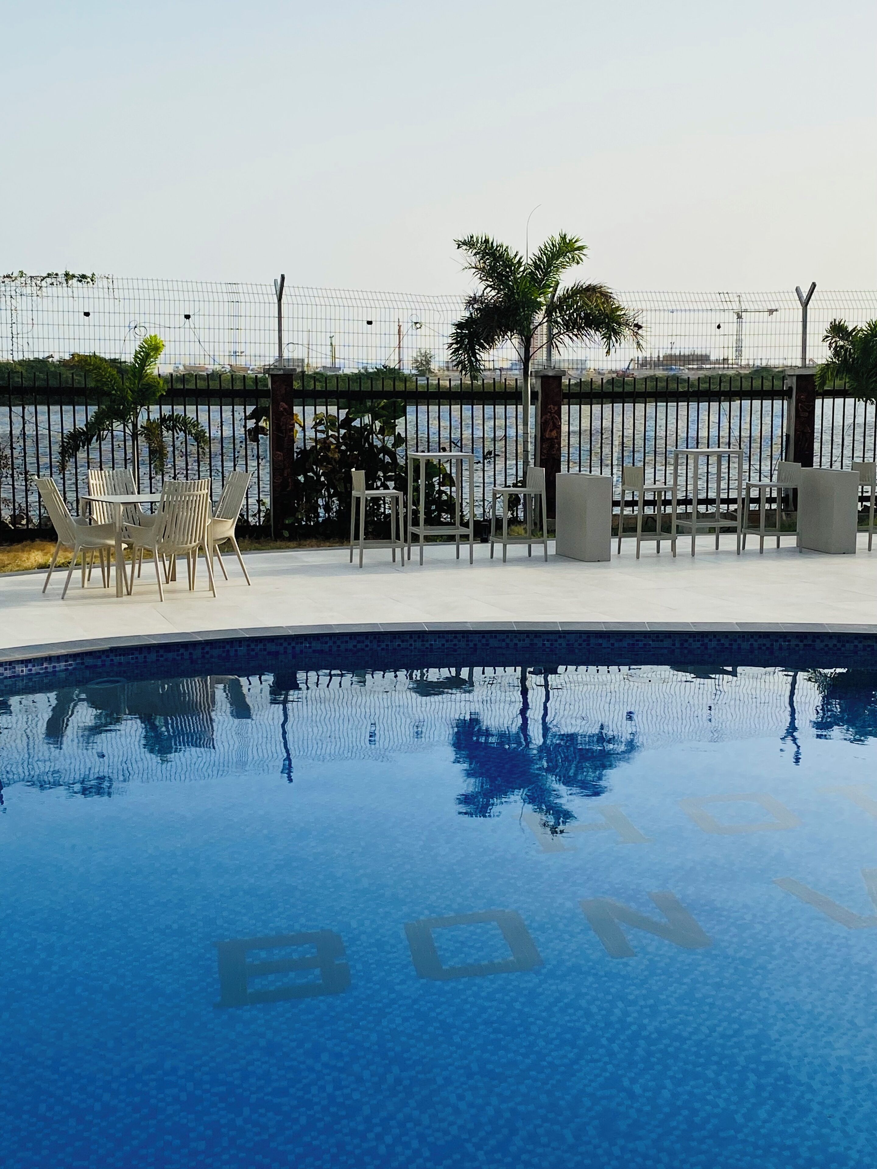 A swimming pool with a clear blue surface, surrounded by a fence and outdoor seating area with chairs and tables.