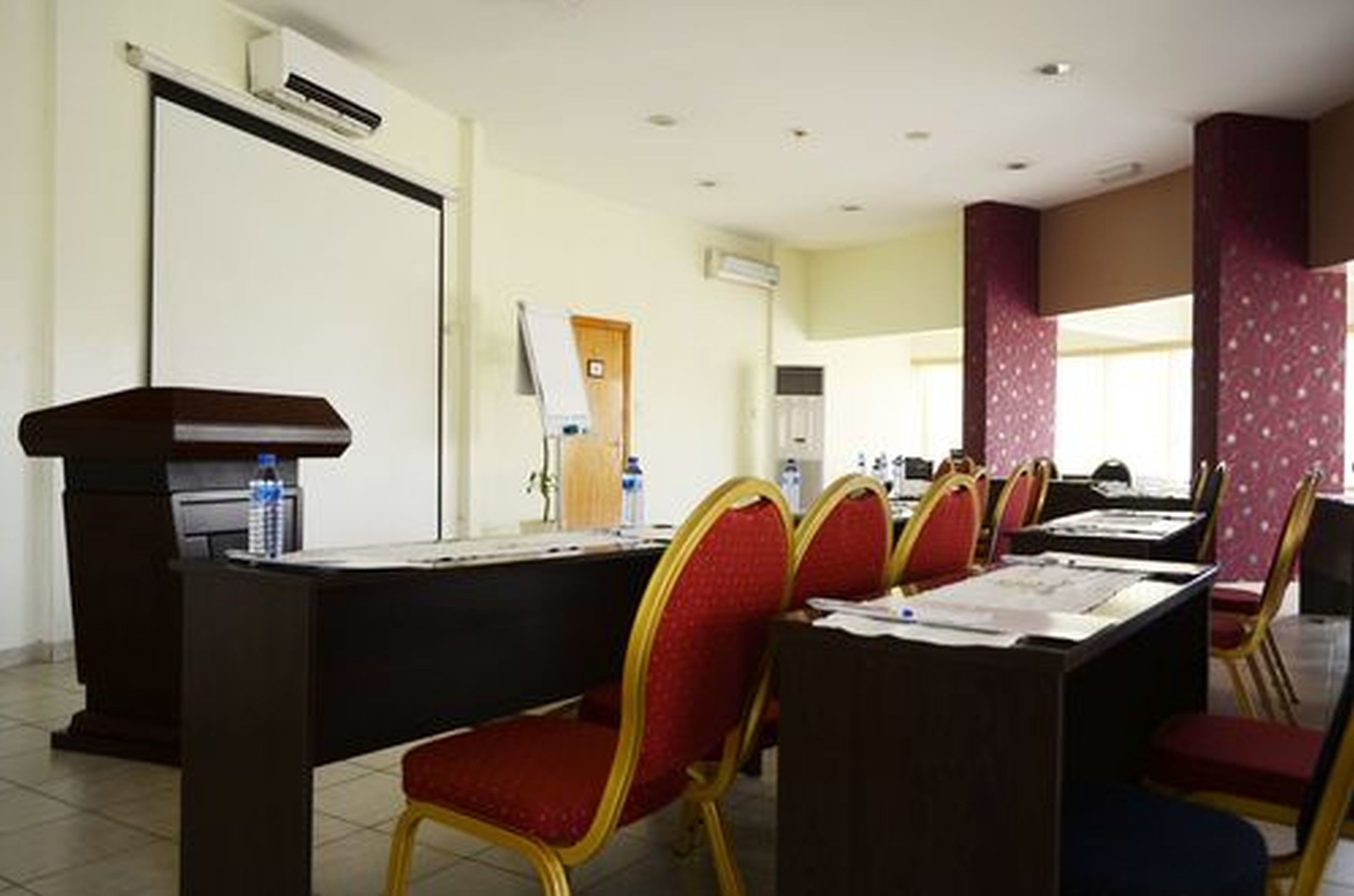 Ein Konferenzraum mit langem Tisch, roten Stühlen, einem Rednerpult und einer Projektionsleinwand.