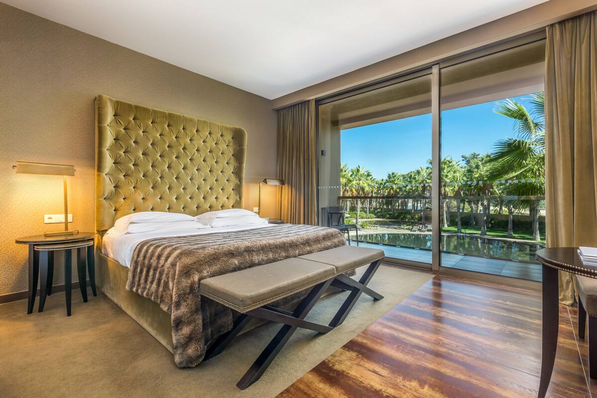 A spacious bedroom with a large bed, a bench, a nightstand, and a view of a pool and palm trees.