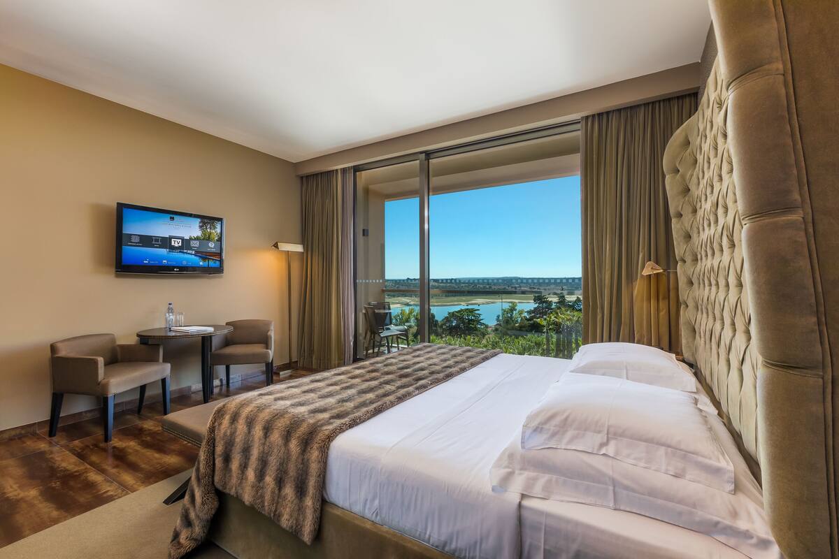 A hotel room with a large bed, a TV mounted on the wall, a desk with chairs, and a view of a body of water through large windows.