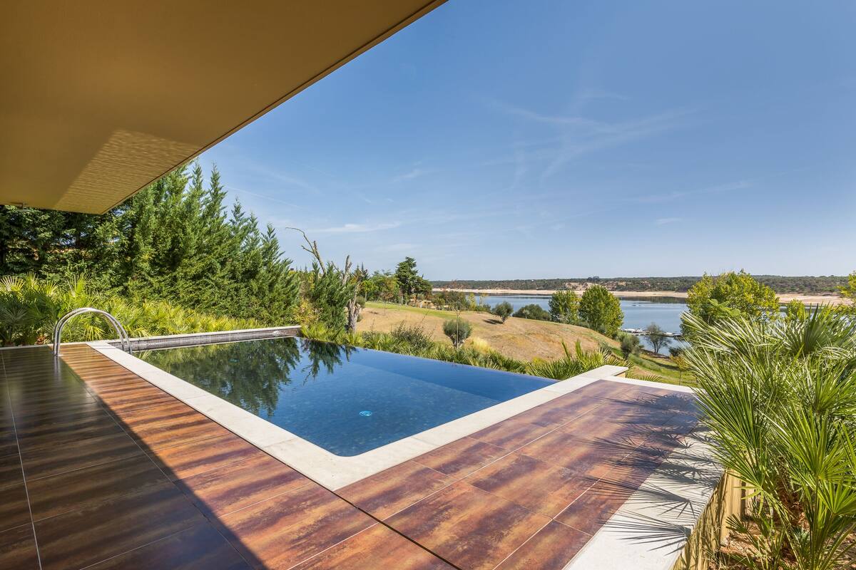 A swimming pool with a wooden deck, surrounded by greenery and a view of a lake and hills.
