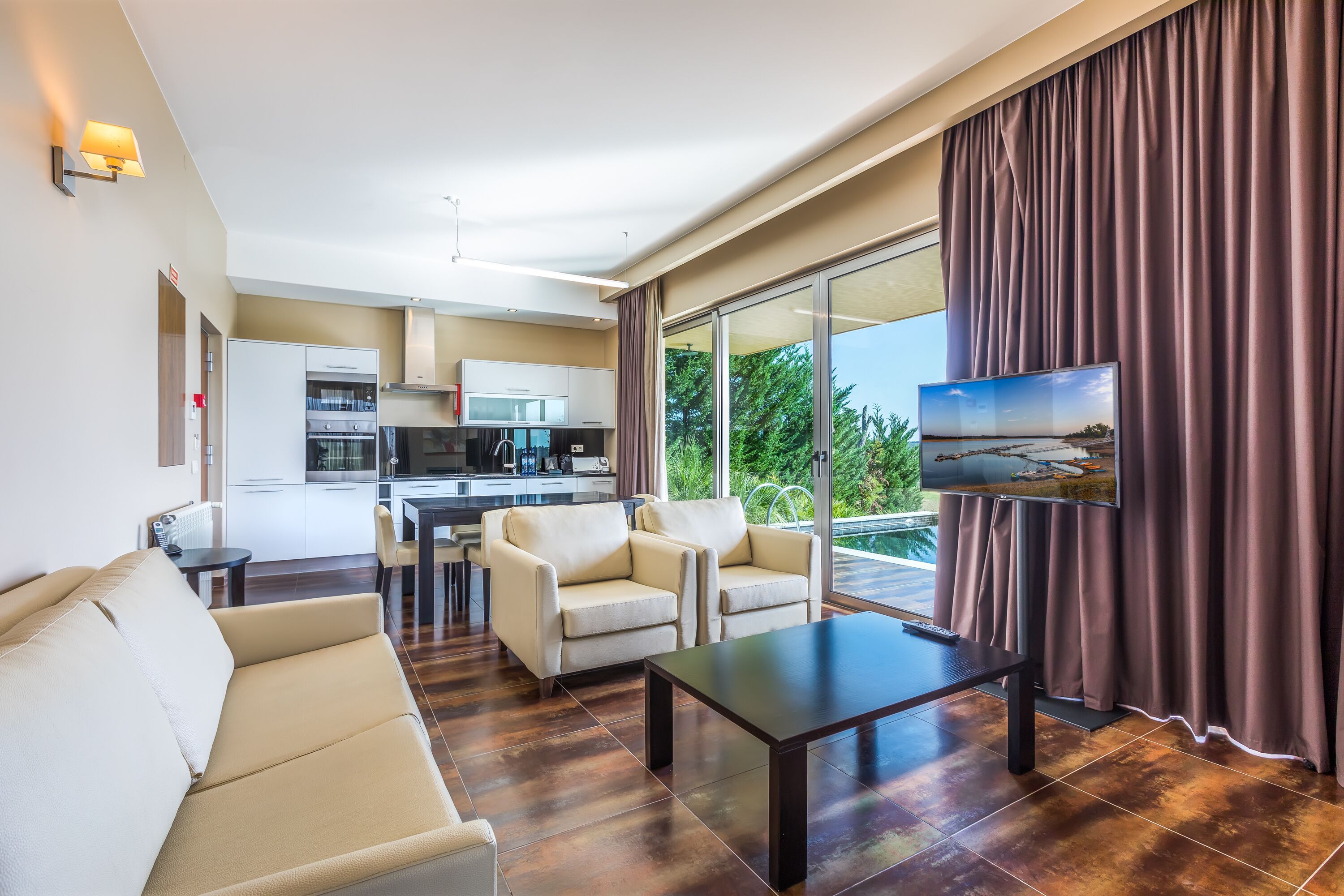 A modern living room with a sofa, armchairs, a coffee table, and a TV. There is a kitchen in the background with a dining area.