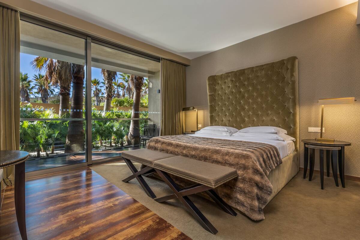A bedroom with a large bed, a bench, a bedside table, and a view of palm trees through a sliding glass door.