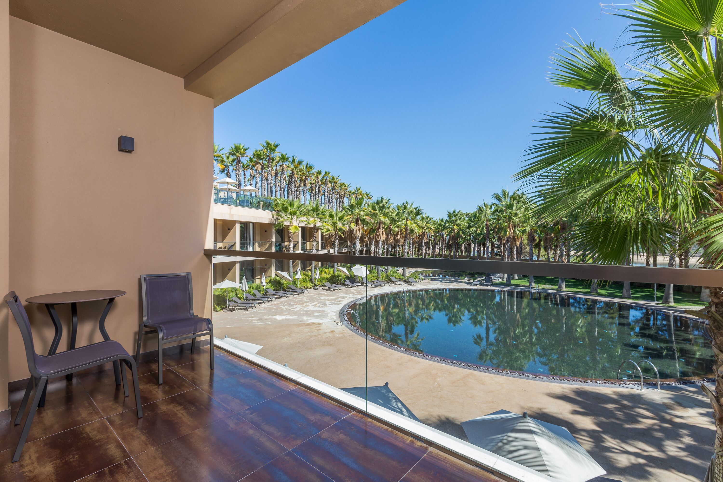A balcony with a pool, chairs, and a view of palm trees and a building.