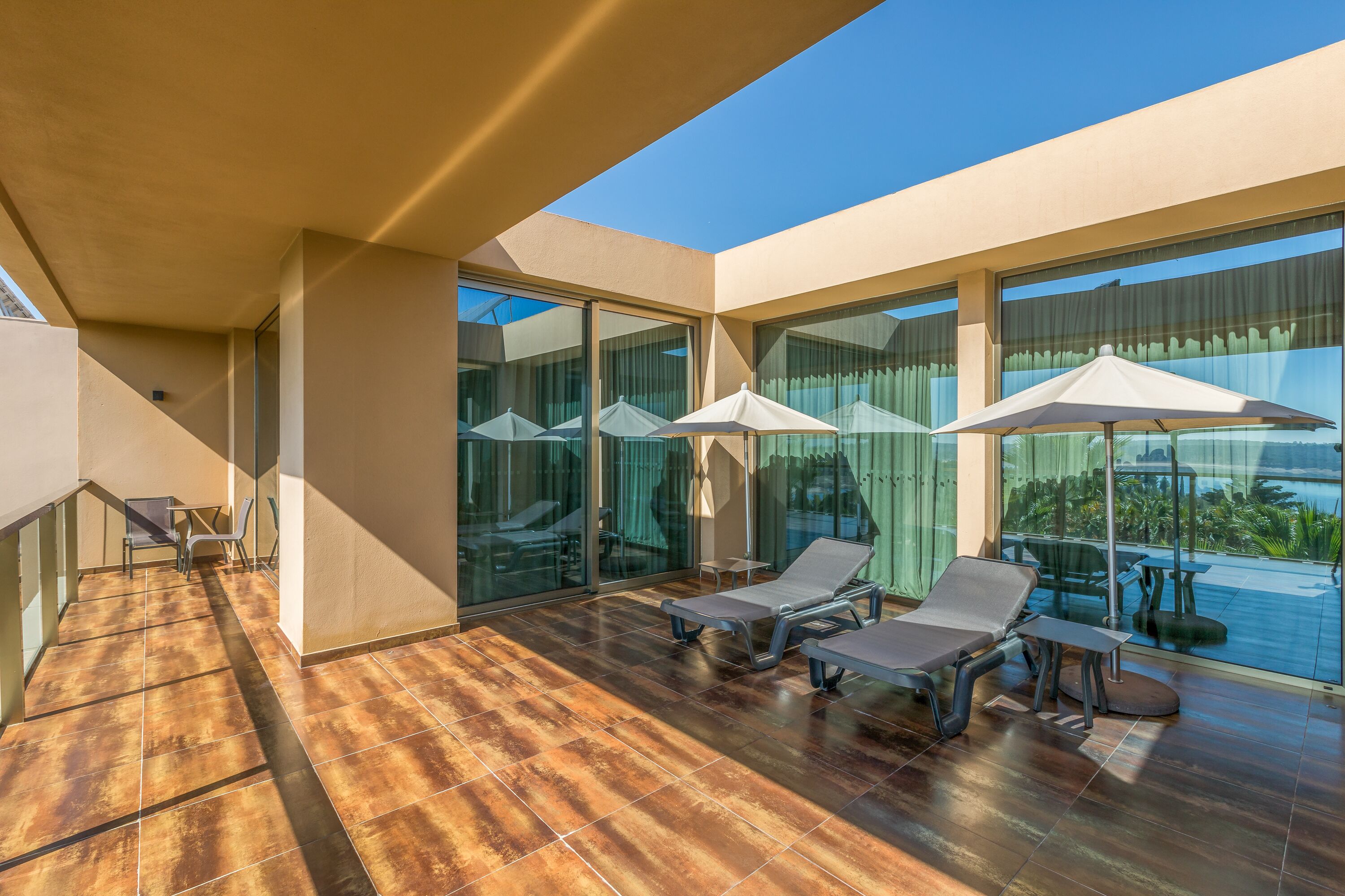 A sunlit balcony with two lounge chairs and umbrellas, large glass windows, and a view of the sea.