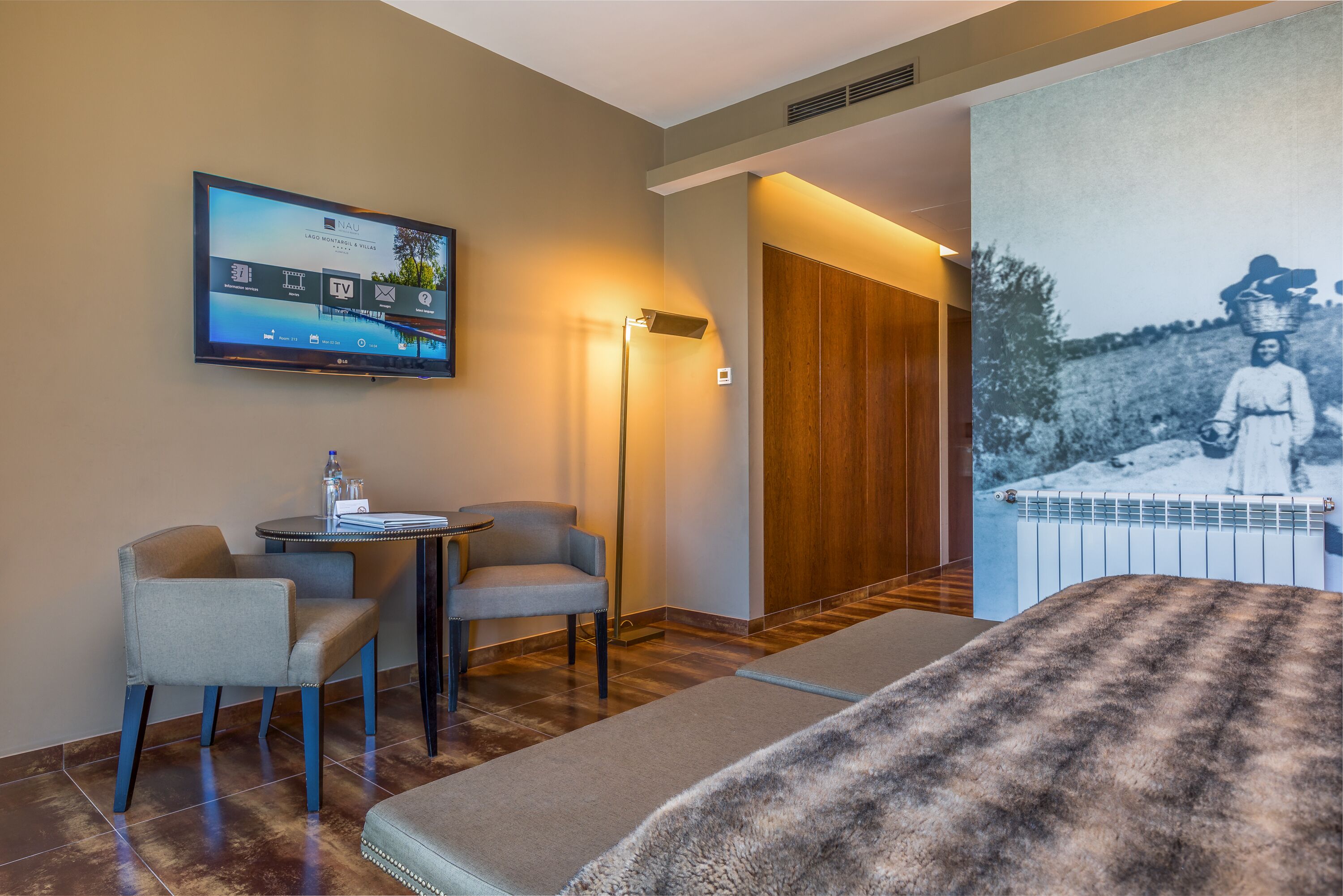 Habitación de hotel con televisor de pantalla plana en la pared, escritorio con dos sillas, ventana grande y puerta de madera.