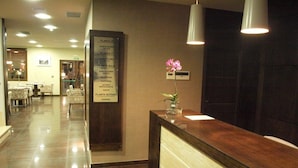 A hotel reception area with a wooden reception desk, a menu board listing various amenities, and a vase with pink flowers. - Hotel Dinastía (Getafe)