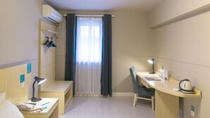 Een kleine hotelkamer met een bed, een bureau met een stoel, een bureaulamp, een waterkoker en een raam met gordijnen.