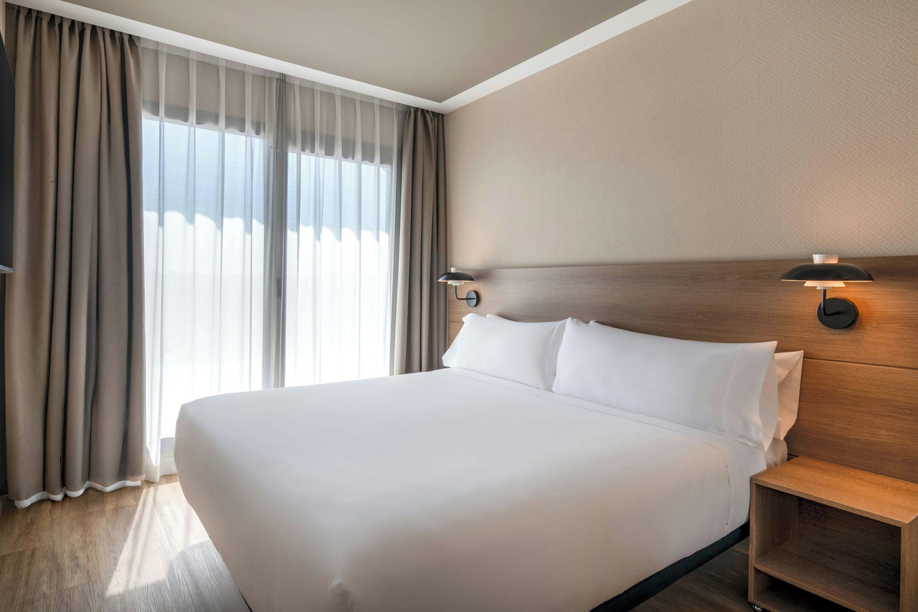 A hotel room with a large bed, a wooden headboard, a nightstand, and a window with curtains.