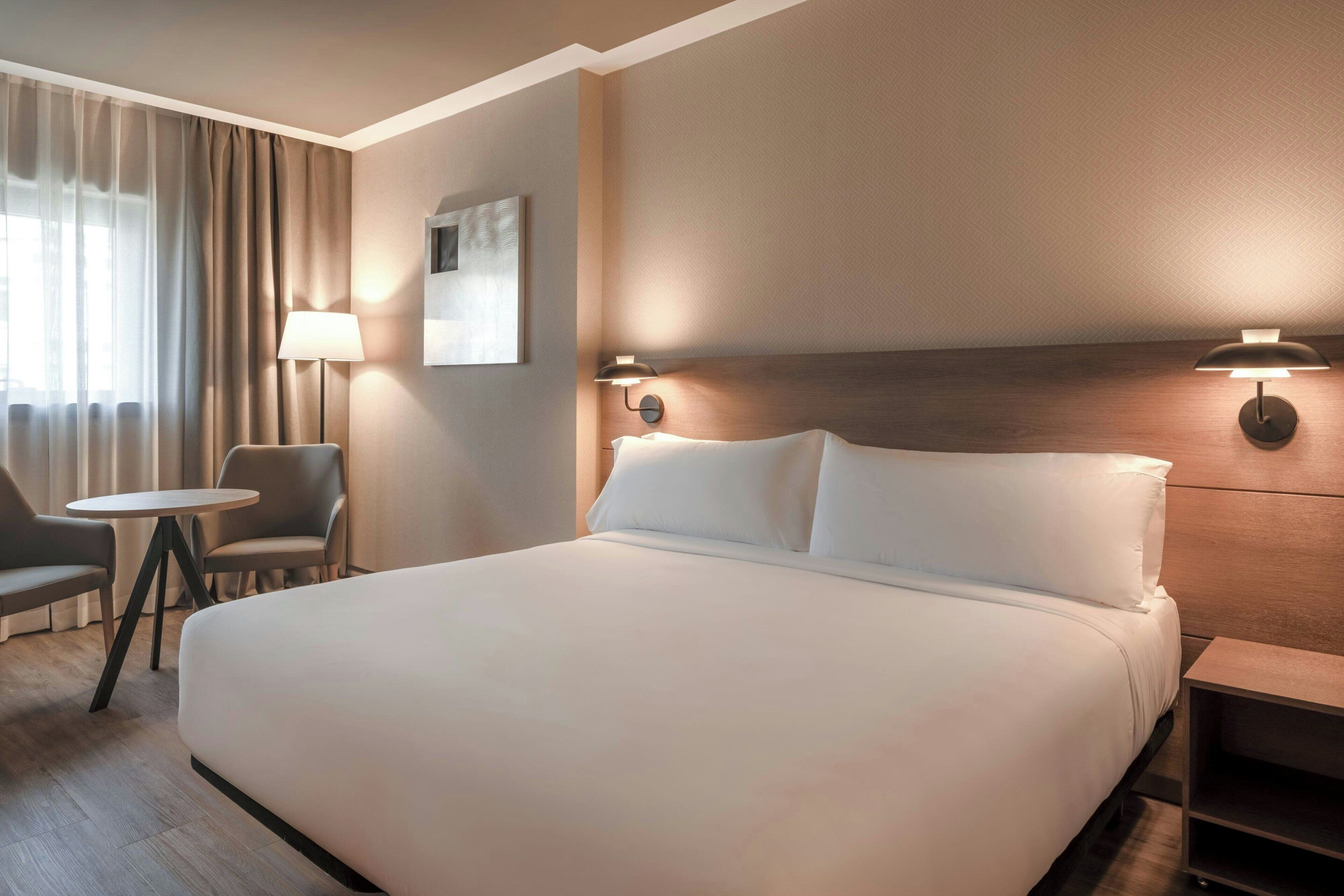 Une chambre d’hôtel avec un grand lit, une table ronde, deux chaises, une lampe et un luminaire fixé au mur.