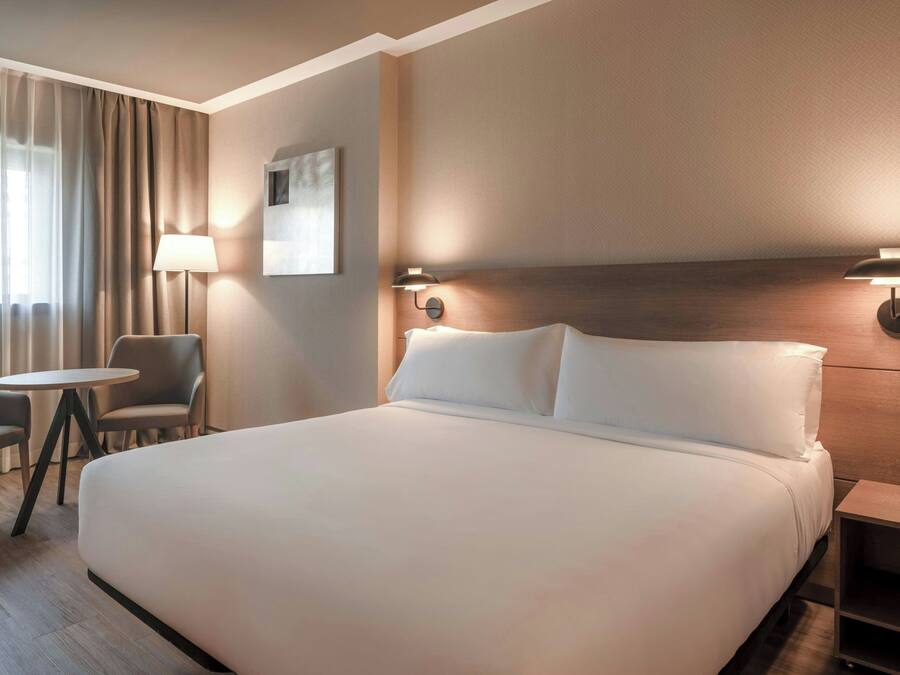 Habitación de hotel con una cama grande, una mesa redonda, dos sillas, una lámpara y un aplique de pared.