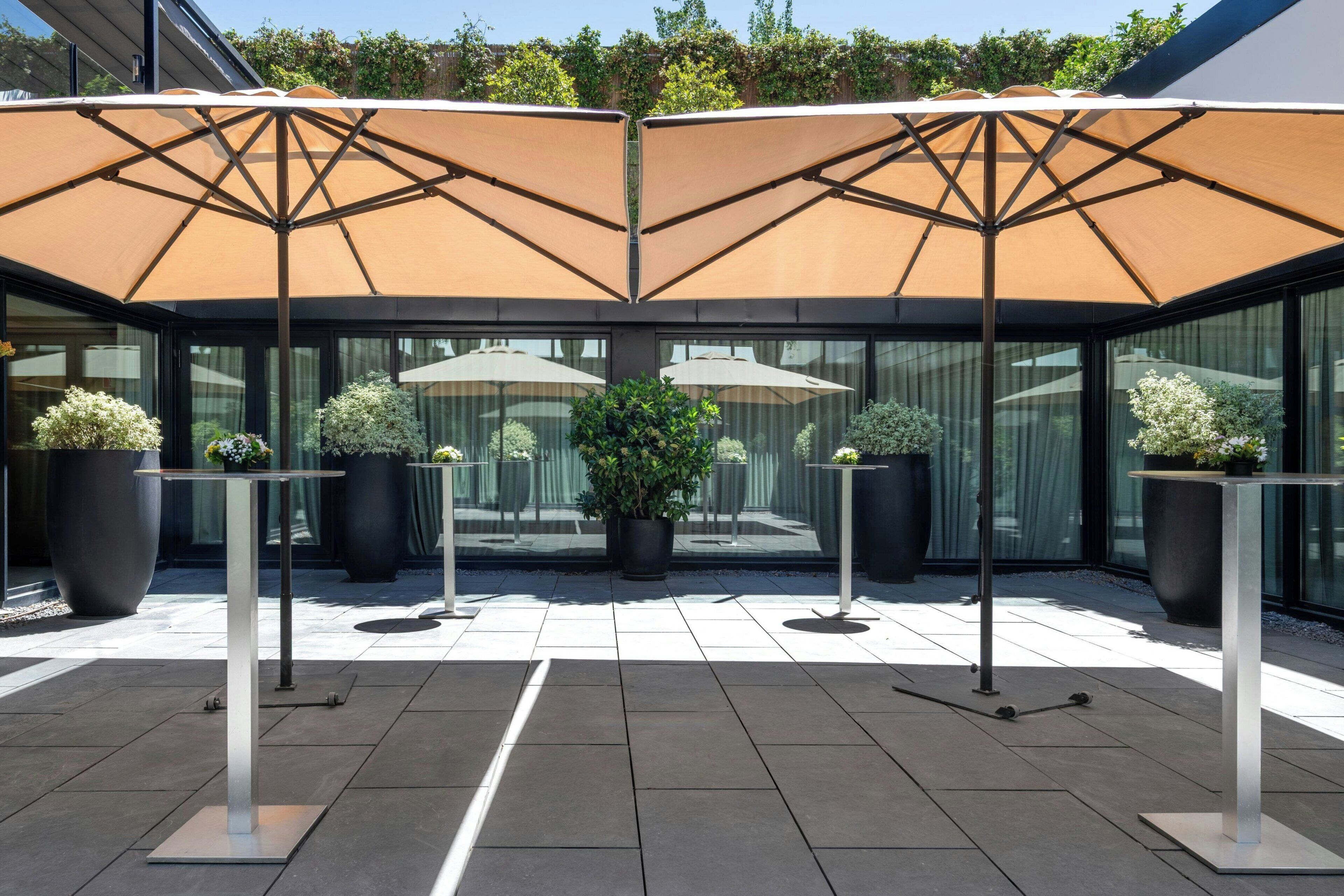 An outdoor patio area with a modern design, featuring a glass enclosure, potted plants, and outdoor seating with umbrellas.
