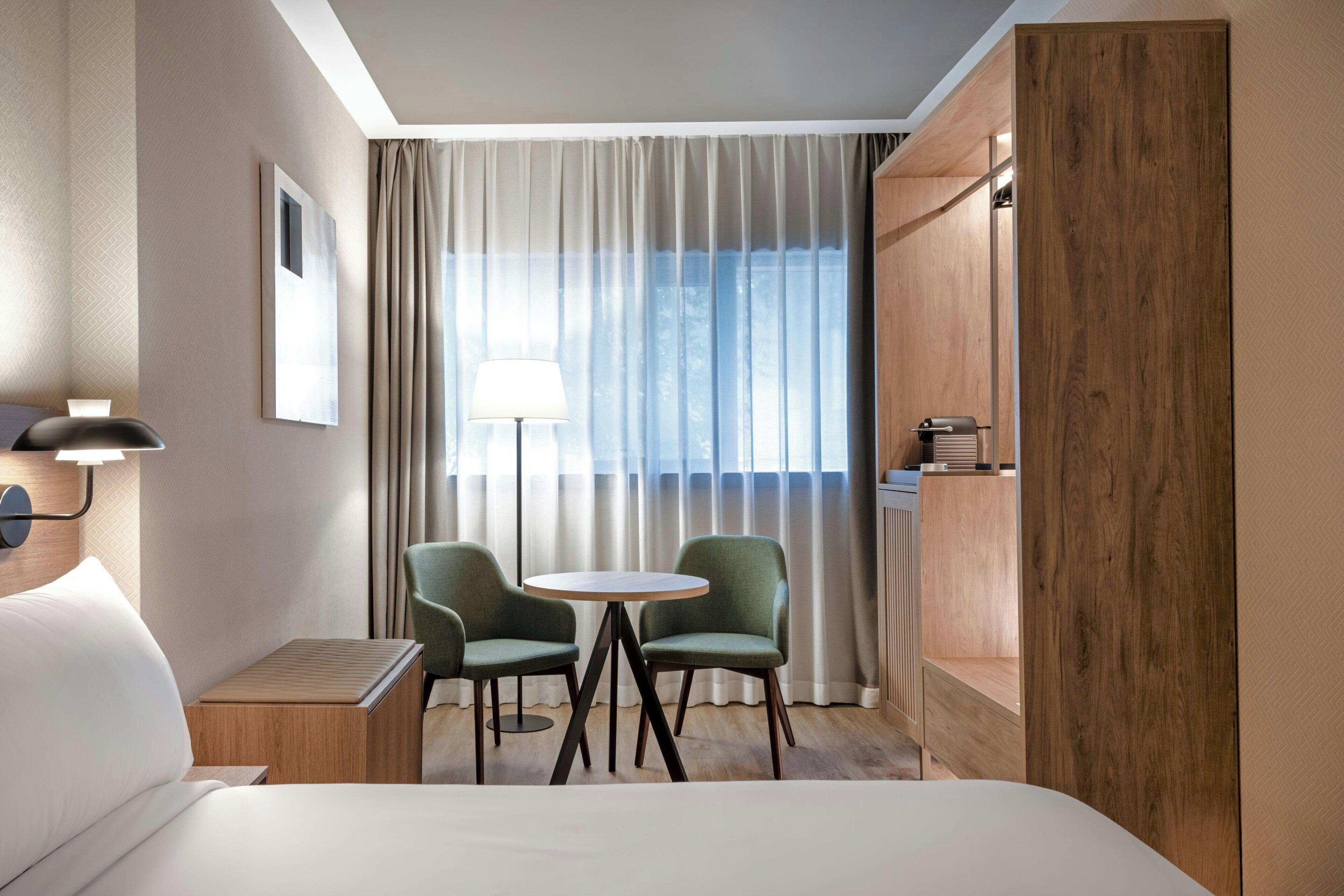 A hotel room with a bed, two armchairs, a small table, a lamp, a window with curtains, and a wooden wardrobe.