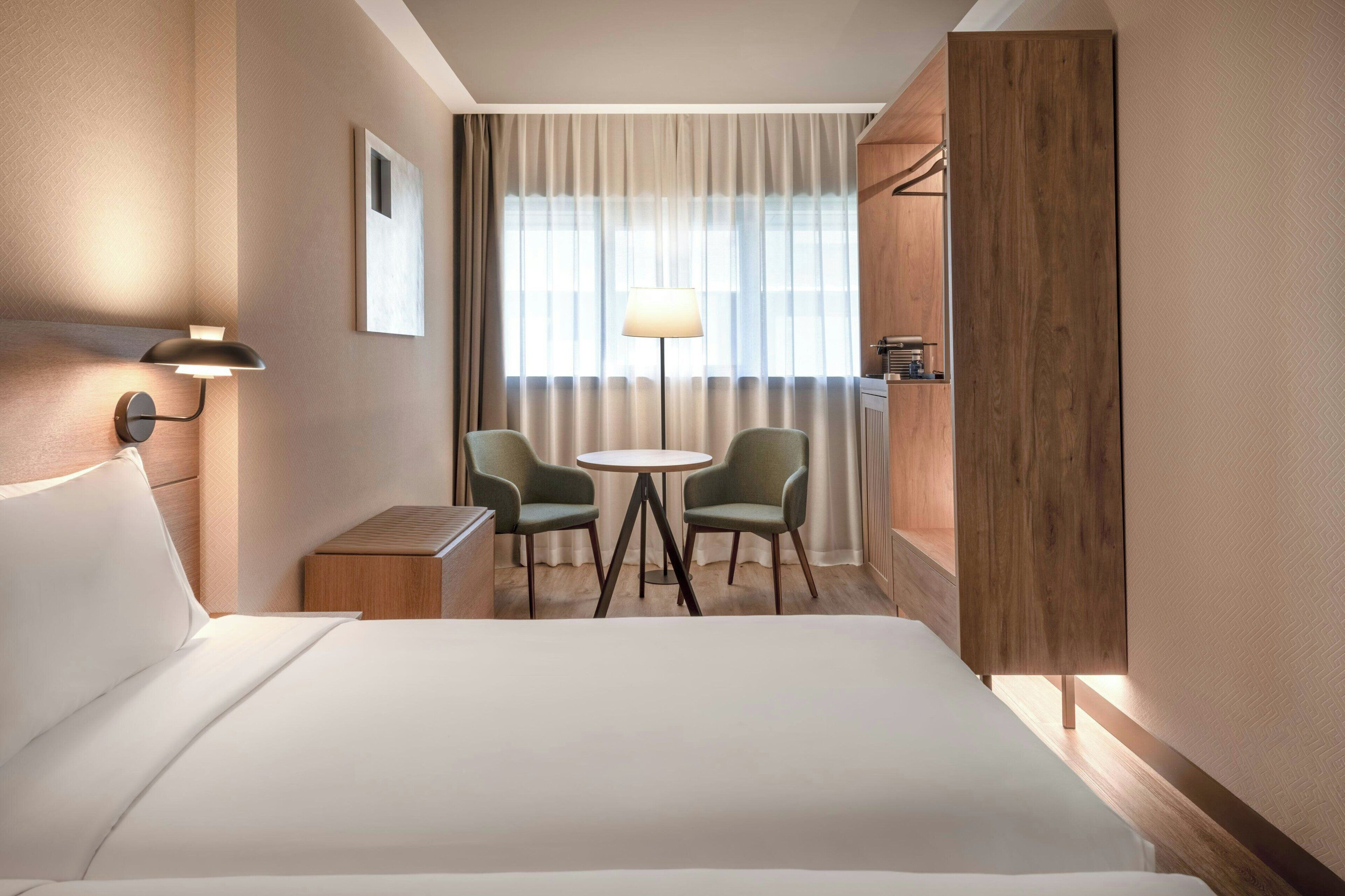 Ein Hotelzimmer mit einem Bett, zwei Stühlen, einem Tisch, einer Lampe und einem Fenster mit Vorhängen.