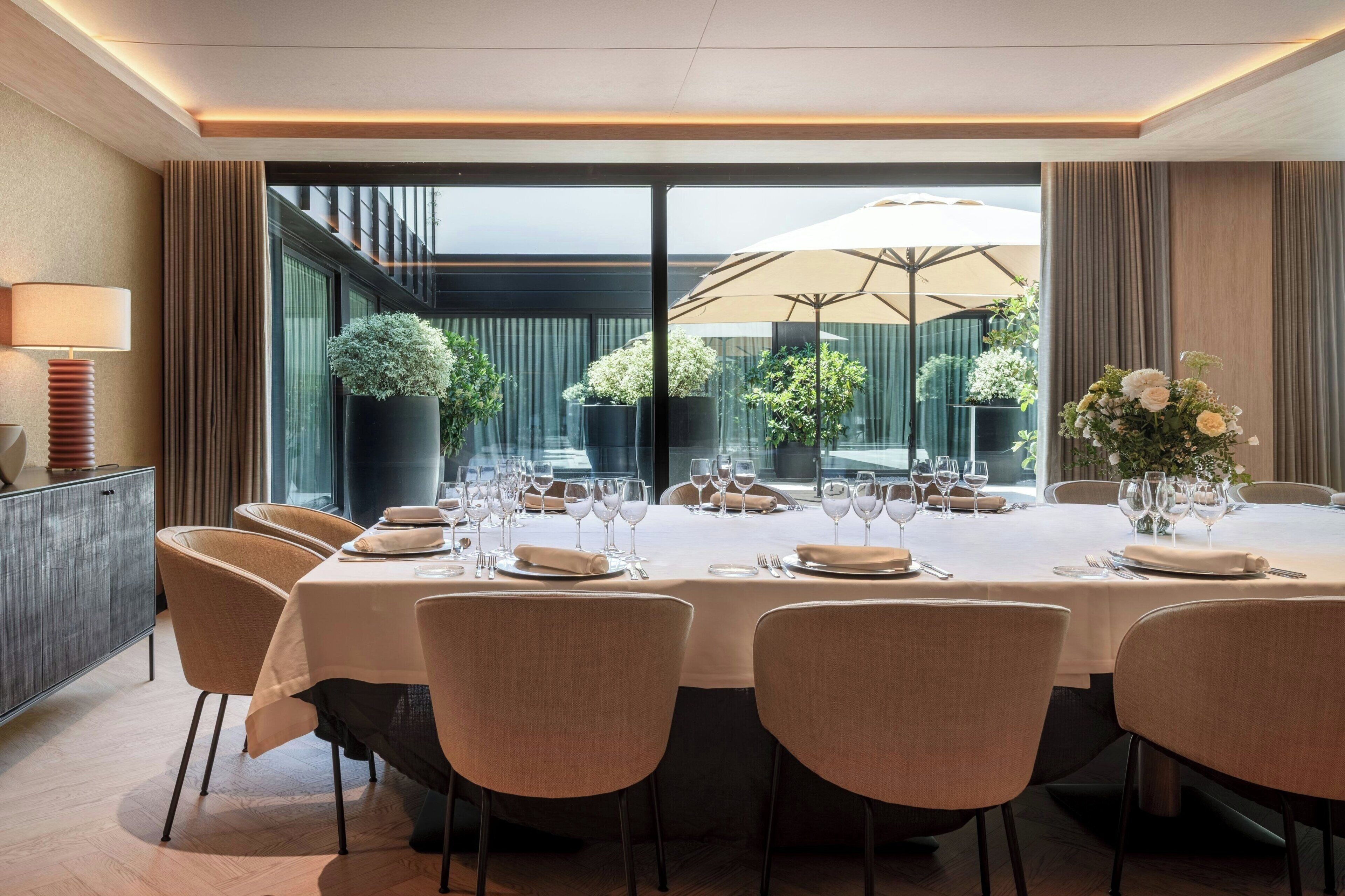 A dining area with a large table set for multiple guests, surrounded by chairs, with a view of greenery outside.