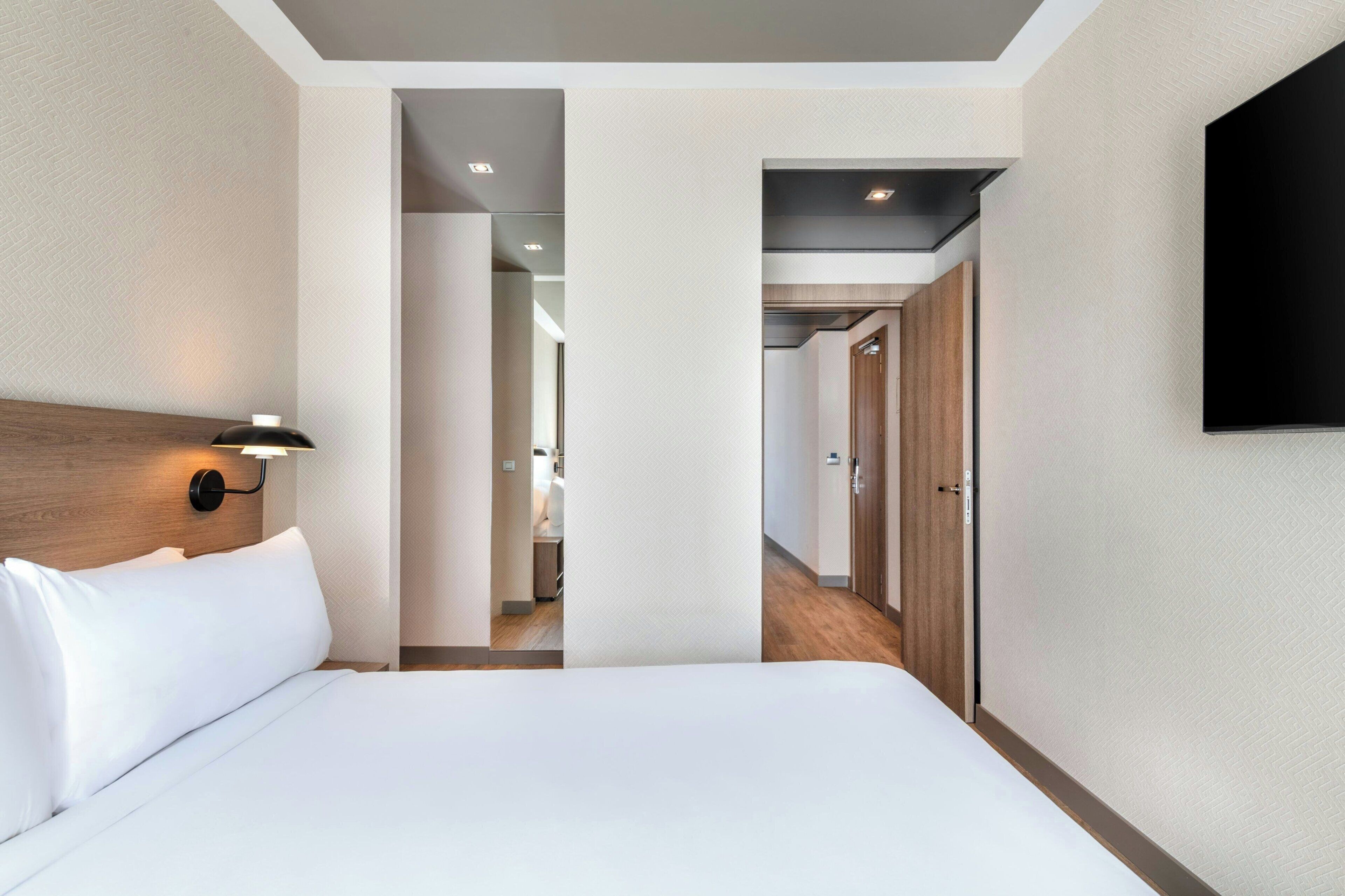 A hotel room with a bed, a wall-mounted TV, and a hallway leading to other rooms.