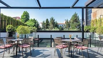 An outdoor patio area with tables and chairs, surrounded by glass walls and a view of trees and buildings.