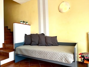 A compact bed with a quilted mattress, a wooden headboard, and a small shelf with plants.