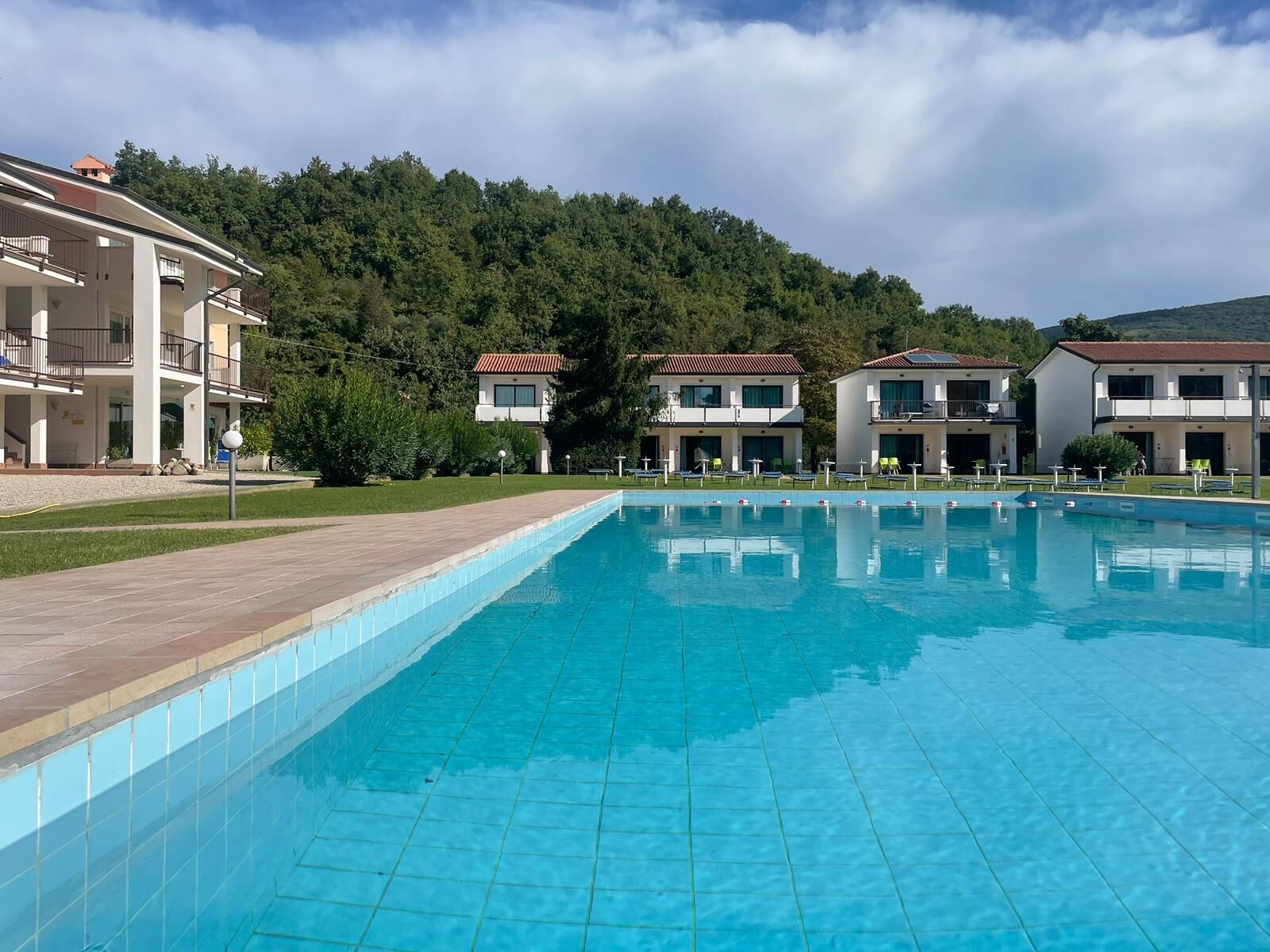 A swimming pool with a tiled edge, surrounded by a paved walkway. There are multiple buildings with balconies and a forested hill in the background.