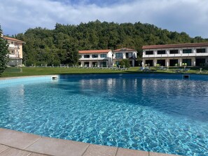 A swimming pool with clear blue water, surrounded by a grassy area and a row of buildings with balconies. - Bran & Denise - Apartments (Costermano sul Garda)