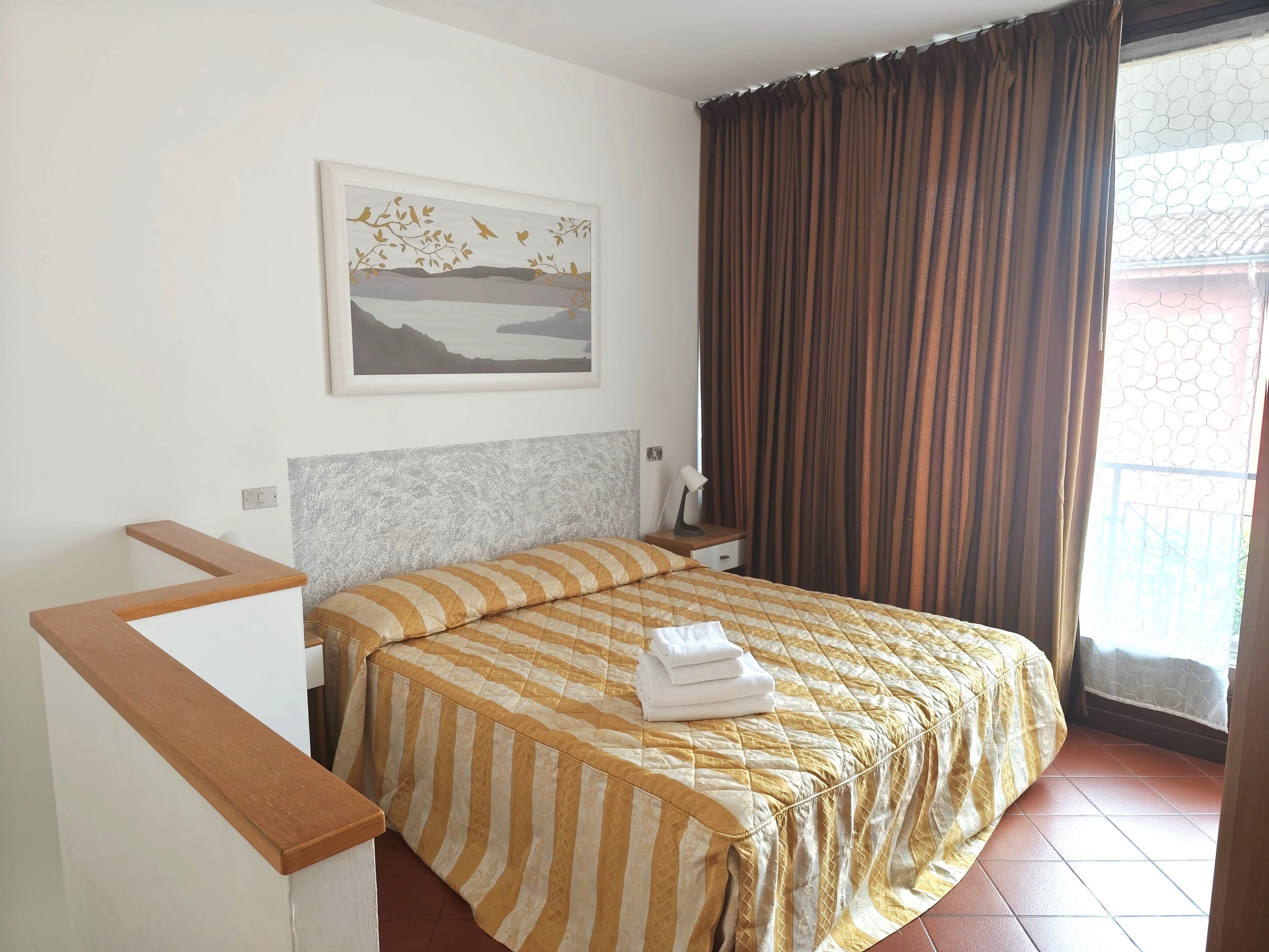 Una habitación de hotel con una cama, una mesita de noche, una lámpara, un cuadro en la pared y una ventana con cortinas.