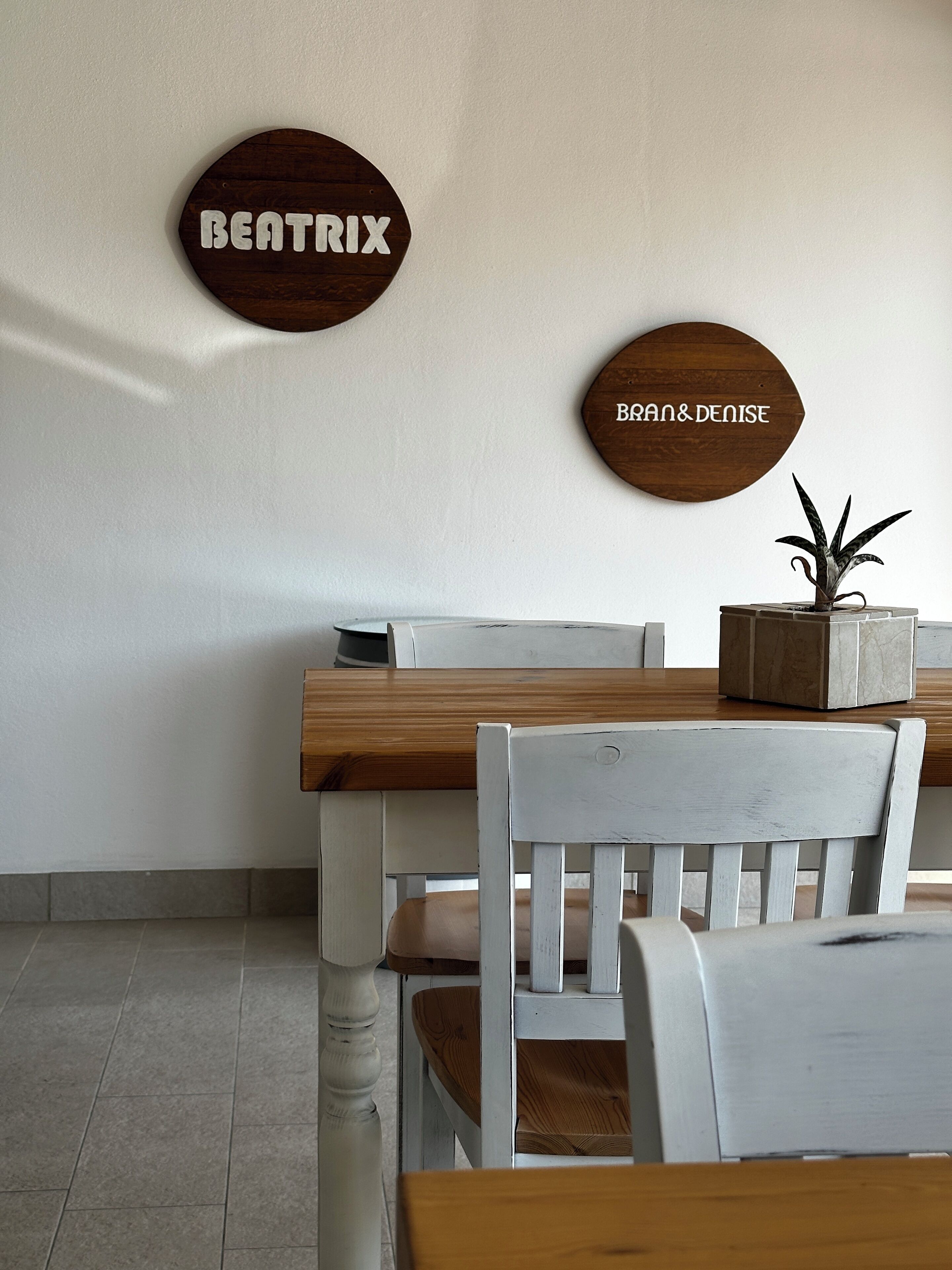 Un área de comedor con una mesa y sillas de madera, una planta en maceta sobre la mesa, y dos letreros de madera en la pared.