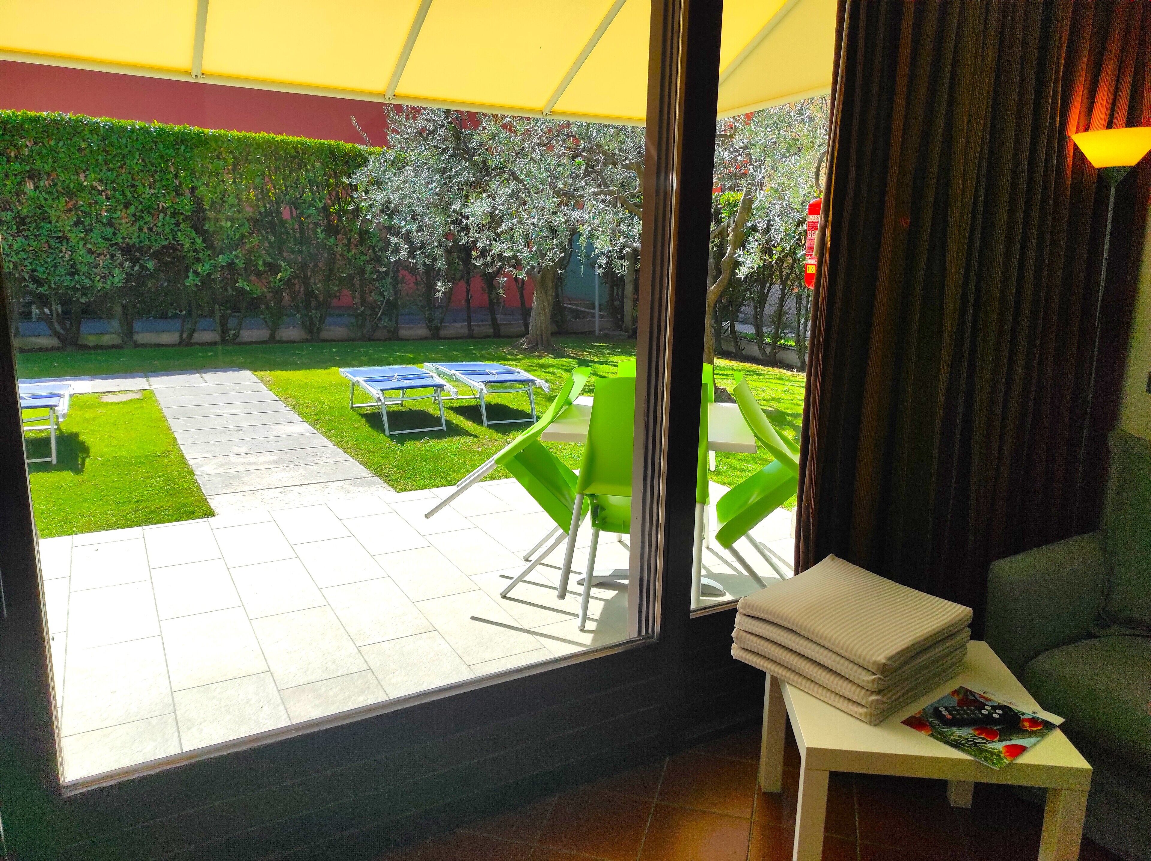 A room with a view of a courtyard featuring a lawn, a table with chairs, and a table with a stack of folded towels.
