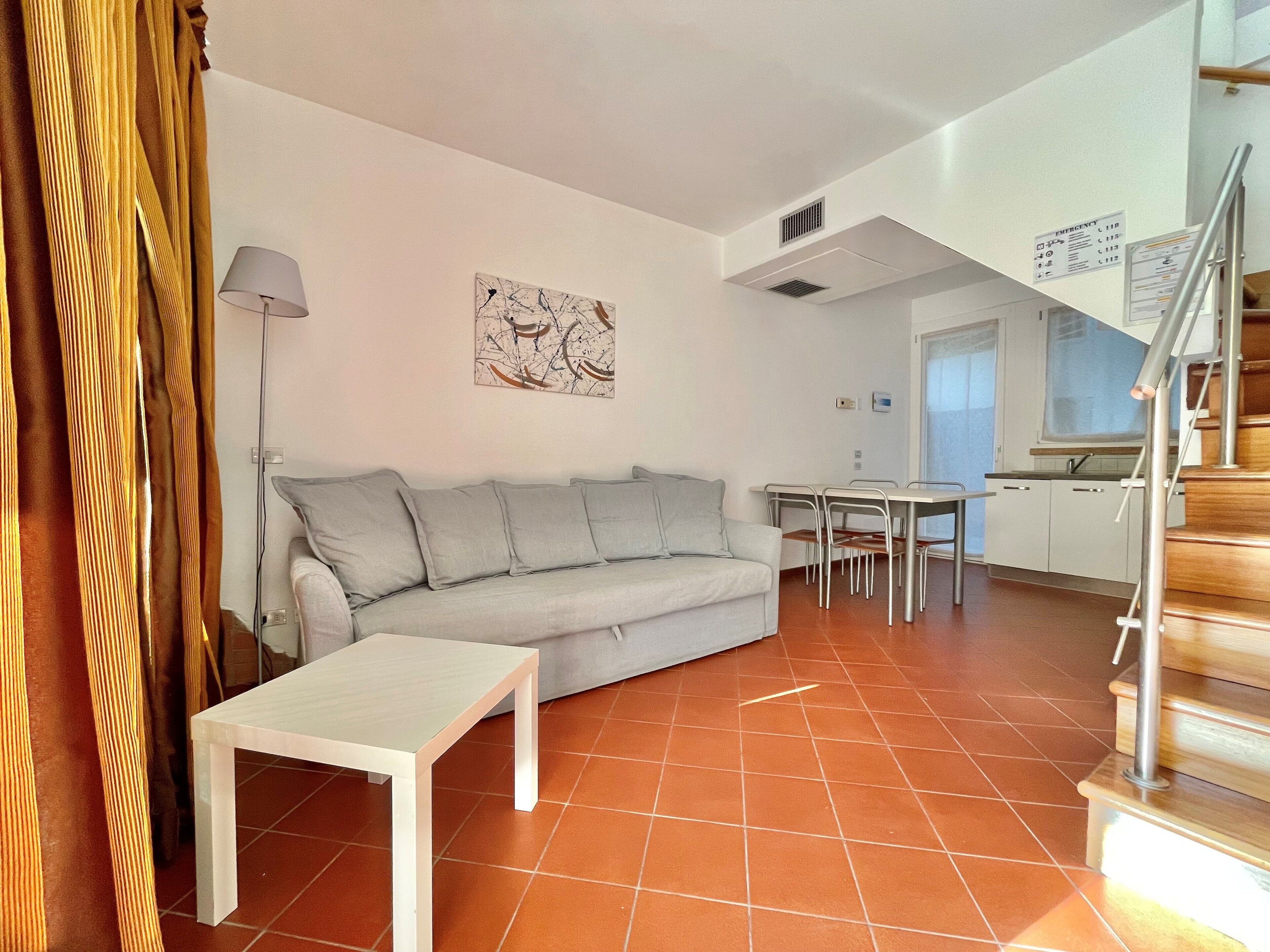 A modern living room with a grey sofa, a small white table, and a dining area with a white table and chairs. There is a staircase with a metal railing leading to an upper level.