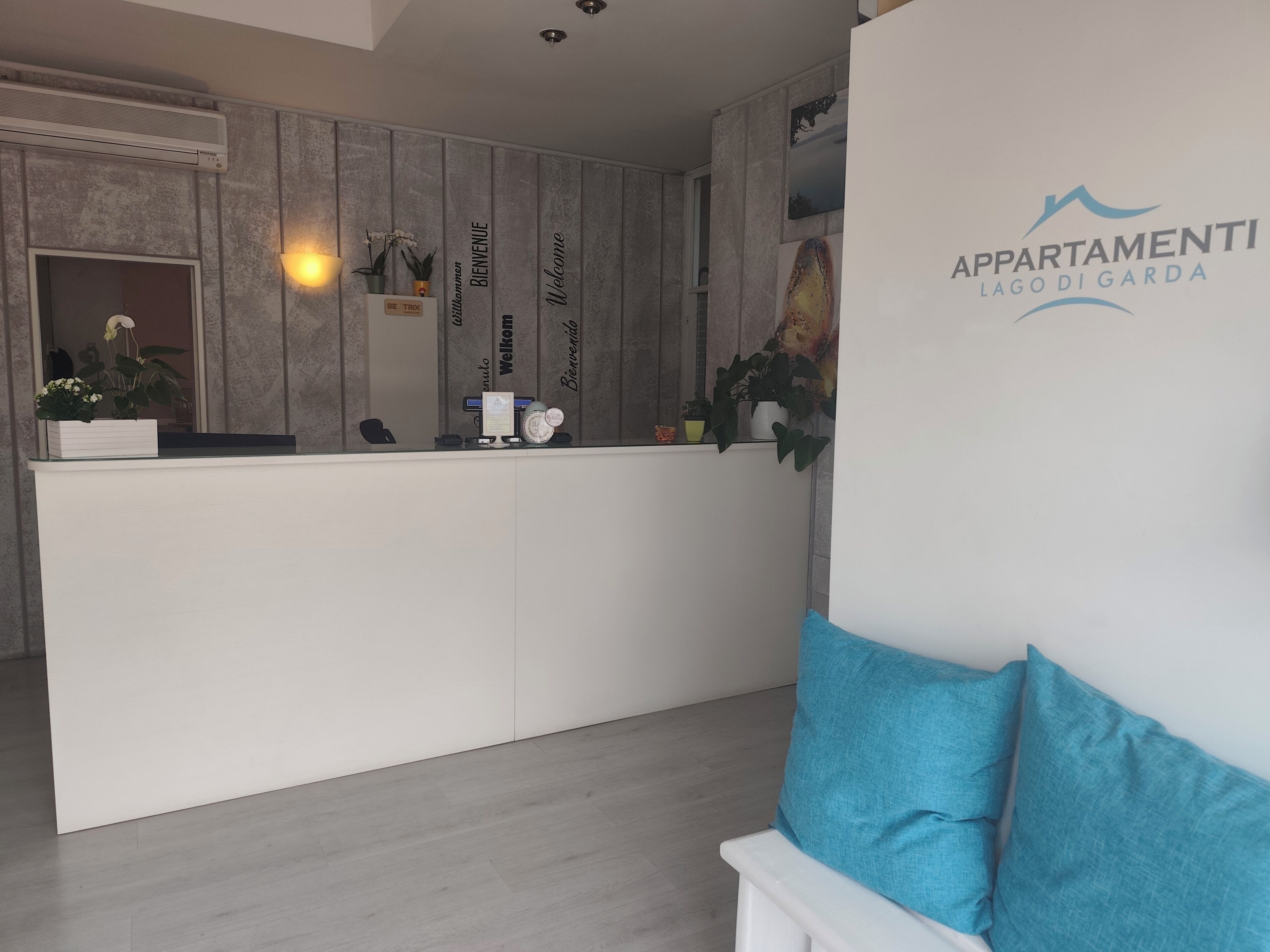 Een receptiebalie met een bord met de tekst 'Welcome', een bord met 'Be Trox' en een bord met 'Bienvenidos'. Er staat een plant op de balie, een luchtverwarmer aan de muur en het logo van 'APPARTAMENTI LAGO DIGARDA'.