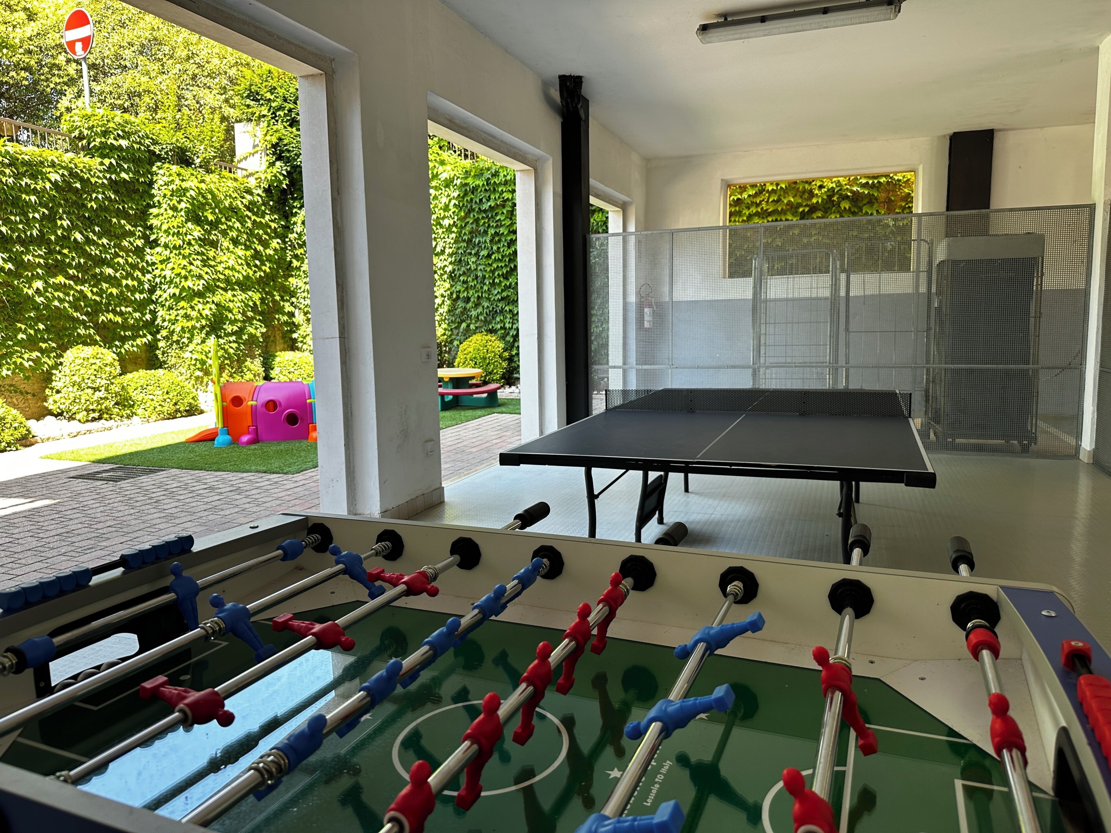 A room with a foosball table, a ping pong table, and a view of a garden with a playground.