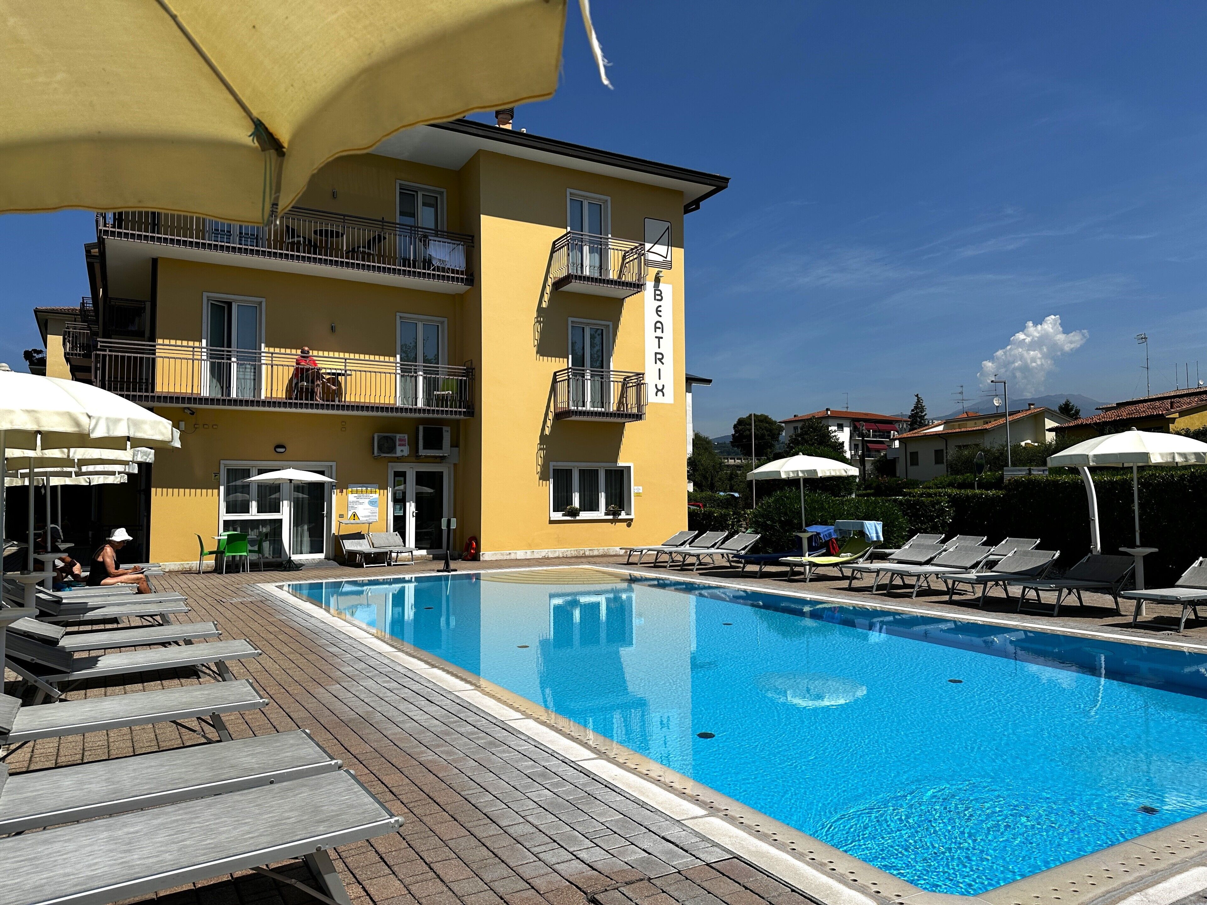 Piscina com espreguiçadeiras e guarda-sóis, um edifício amarelo com varandas e um céu azul límpido.