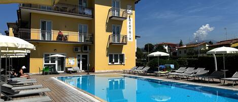 Une piscine avec des chaises longues et des parasols, un bâtiment jaune doté de balcons, et un ciel d’un bleu limpide.