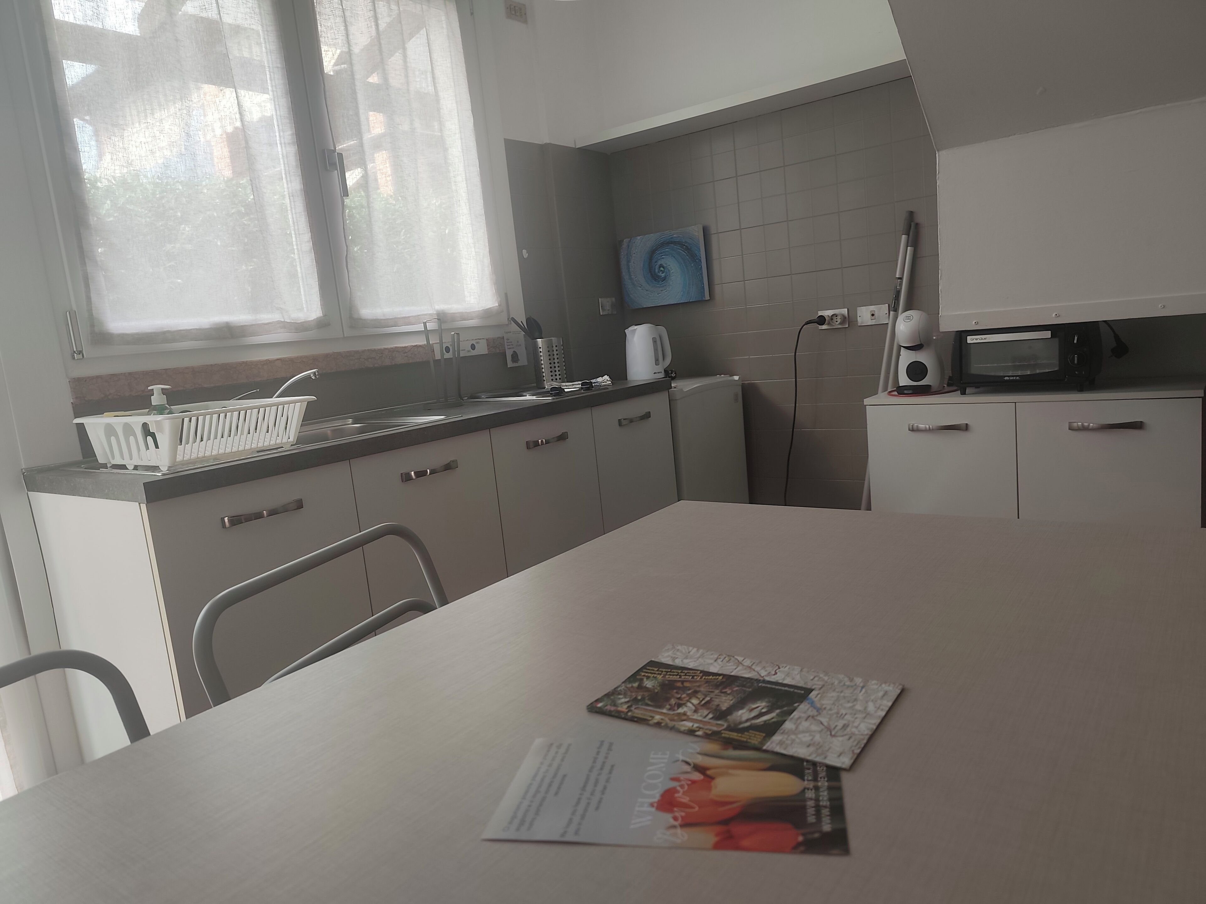A modern kitchen with a white countertop, a kettle, a microwave, and a sink. There is a window with sheer curtains and a painting on the wall.