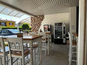 Un café en plein air avec des tables et des chaises en bois peintes en blanc, une voiture garée à l’extérieur et un distributeur automatique.