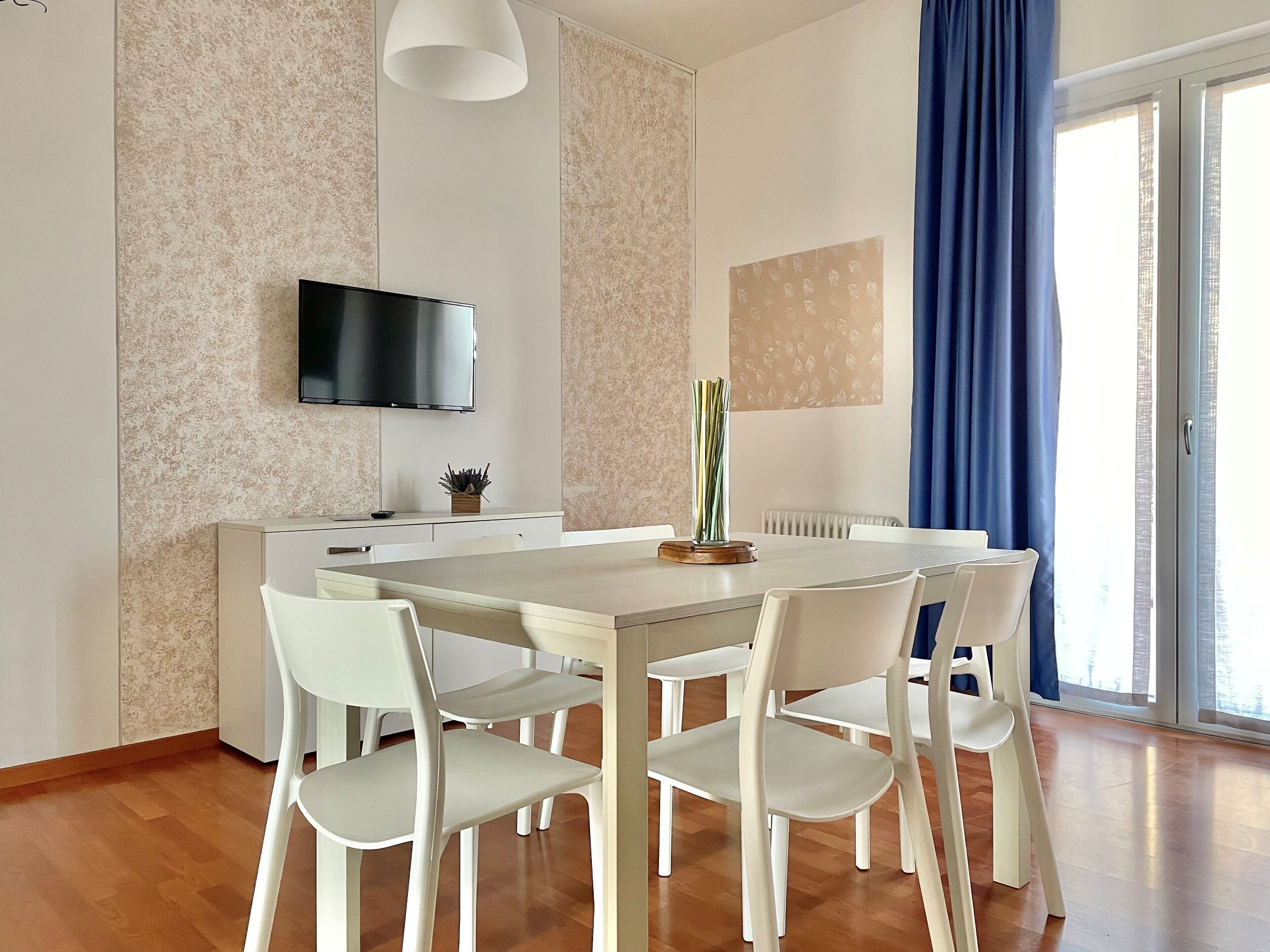 A modern dining area with a white table and chairs, a flat-screen TV mounted on the wall, and a large window with blue curtains.