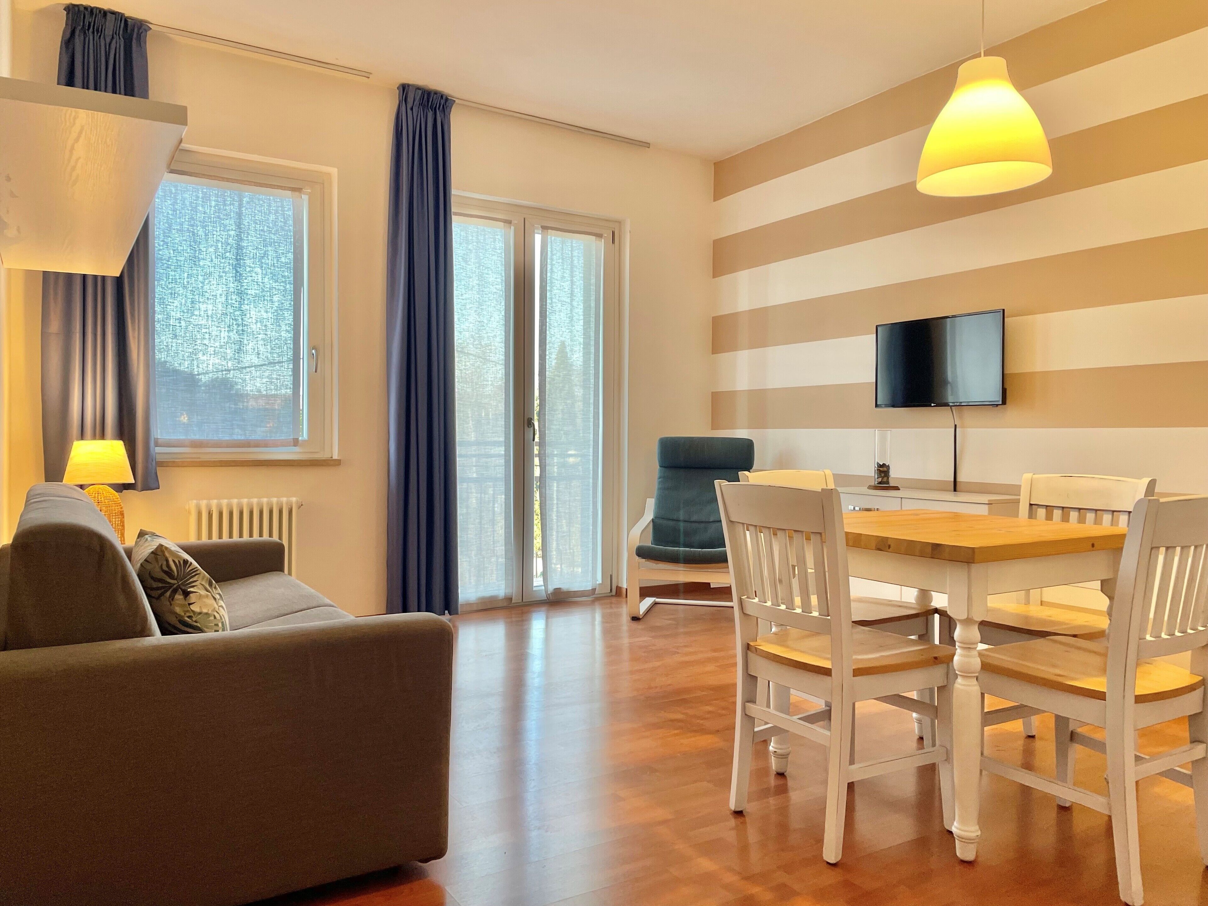 A modern living room with a sofa, a dining table with chairs, a TV mounted on the wall, and large windows with curtains.