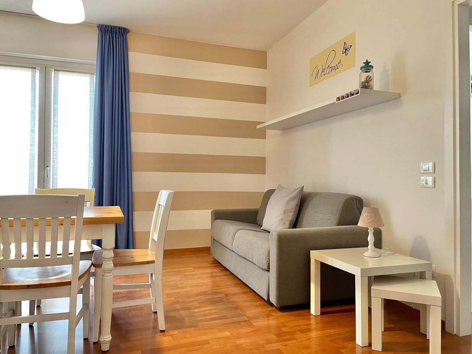 A living room with a sofa, a dining table with chairs, a lamp, and a wall-mounted shelf with decorative items.