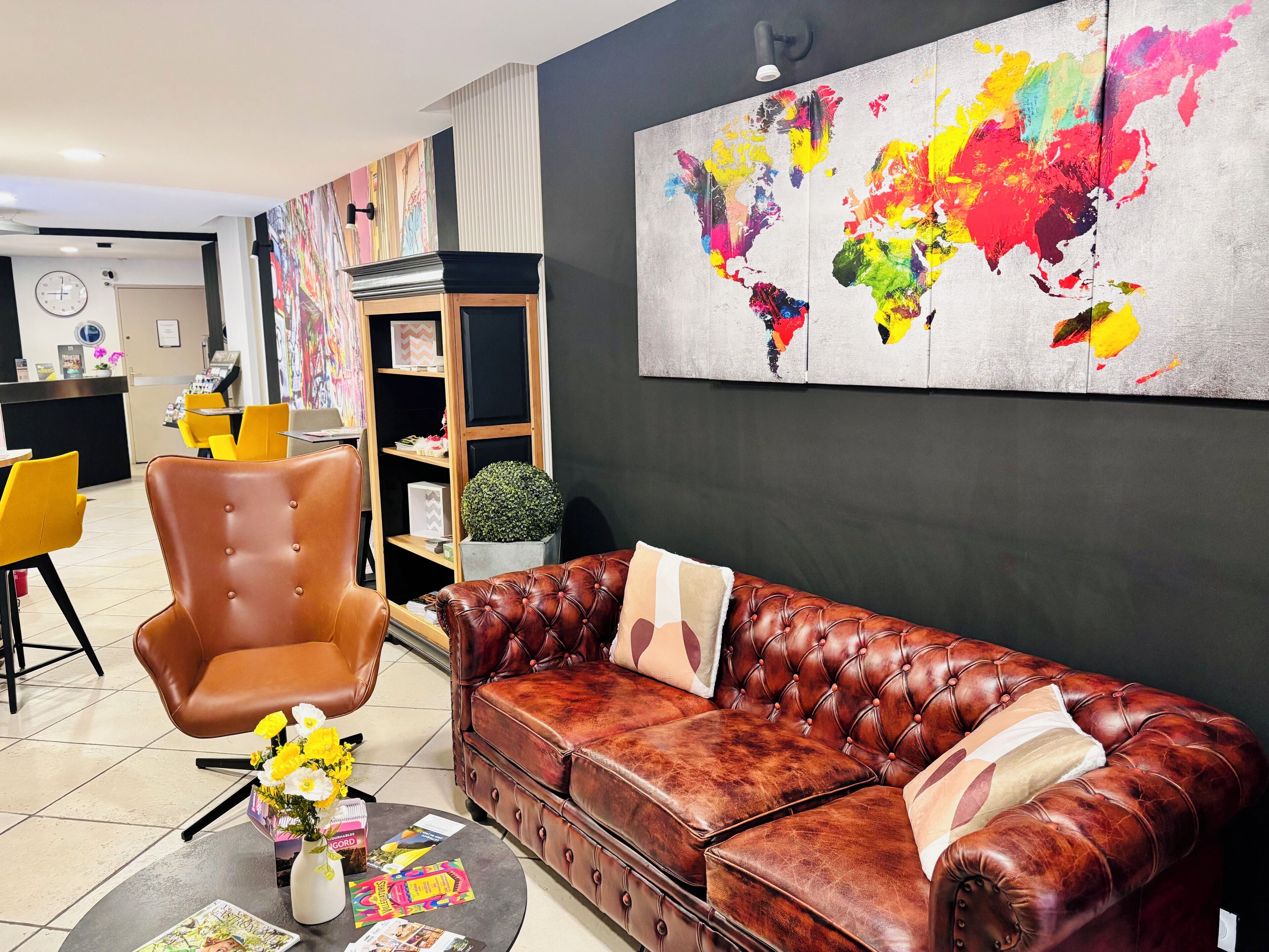 Un salon moderne avec un canapé en cuir, un fauteuil marron et une table basse sur laquelle sont posés des magazines et un vase de fleurs. Un grand tableau mural représentant une carte du monde aux couleurs vives orne le mur.