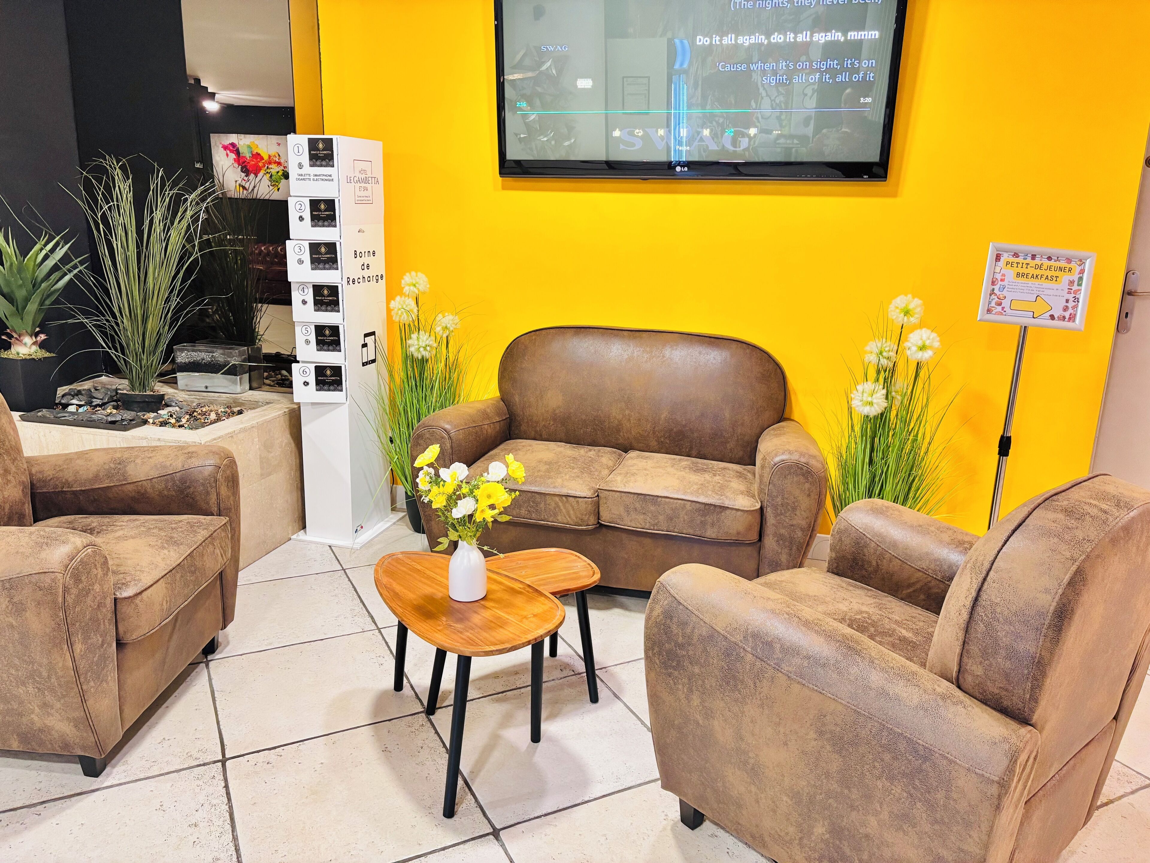 Un área de recepción con un sofá de cuero marrón, una mesa de centro de madera y un jarrón con flores. Hay un televisor en la pared mostrando un programa.