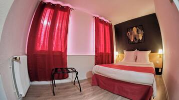 A compact hotel room with a bed, red curtains, a radiator, a small table, and a wall-mounted artwork.