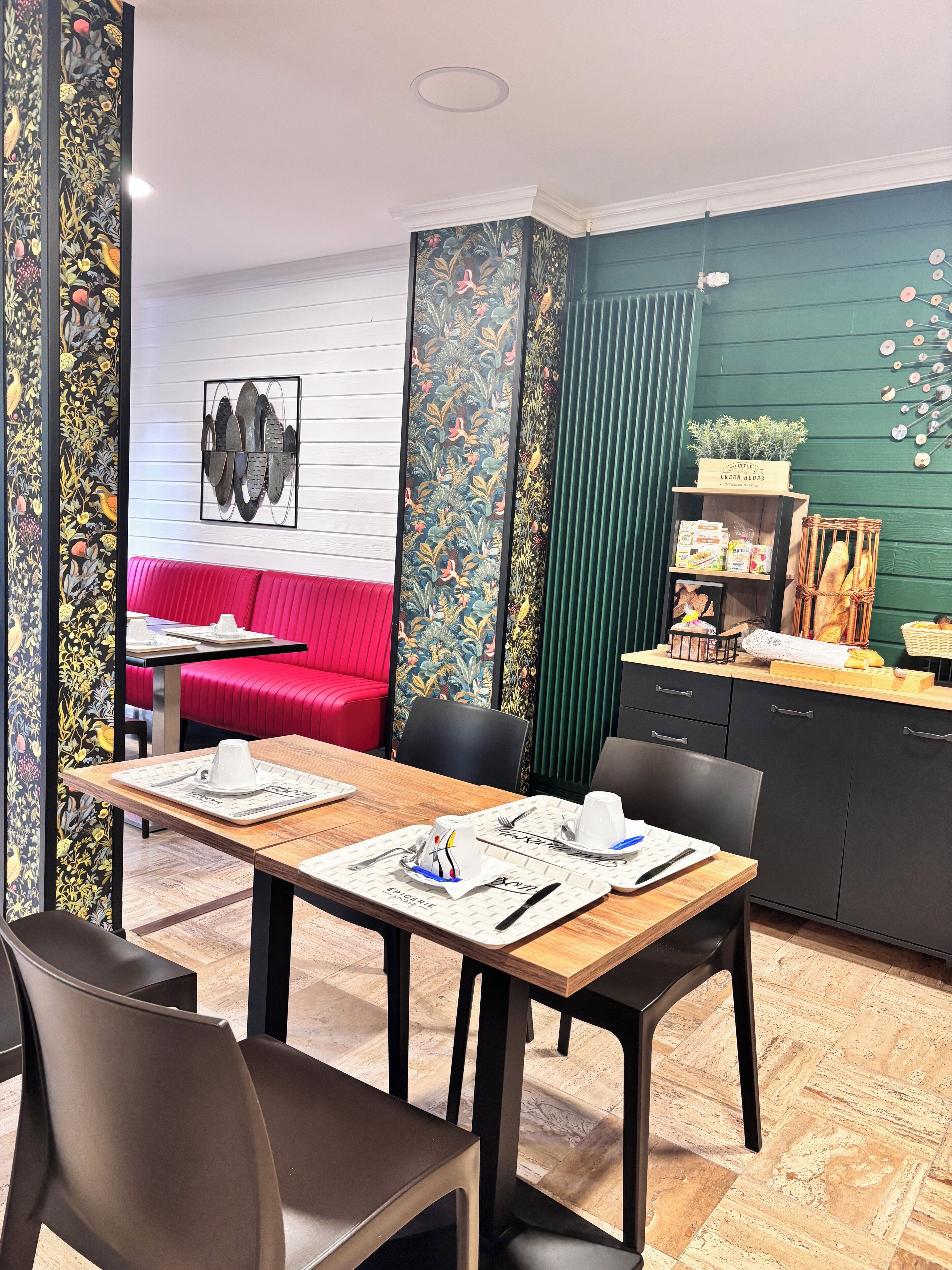 A dining area with a wooden table set for two, a red sofa in the background, and a green wall with a clock.
