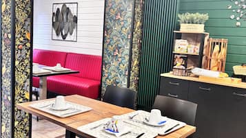 A dining area with a wooden table set for two, a red sofa in the background, and a green wall with a clock.