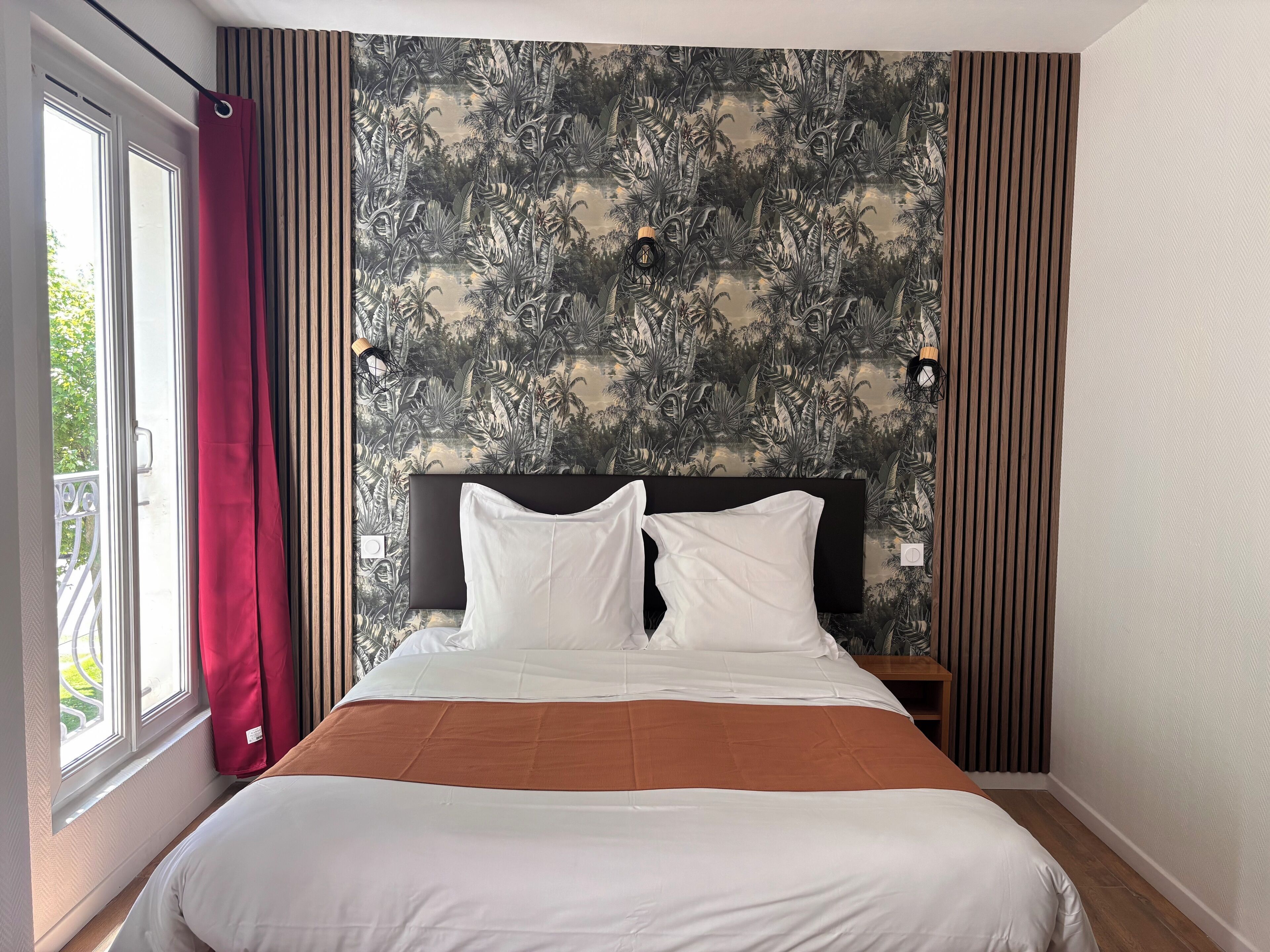 Une chambre avec un grand lit, une fenêtre avec des rideaux, un mur orné d’un motif de feuilles tropicales et deux lampes fixées au mur.
