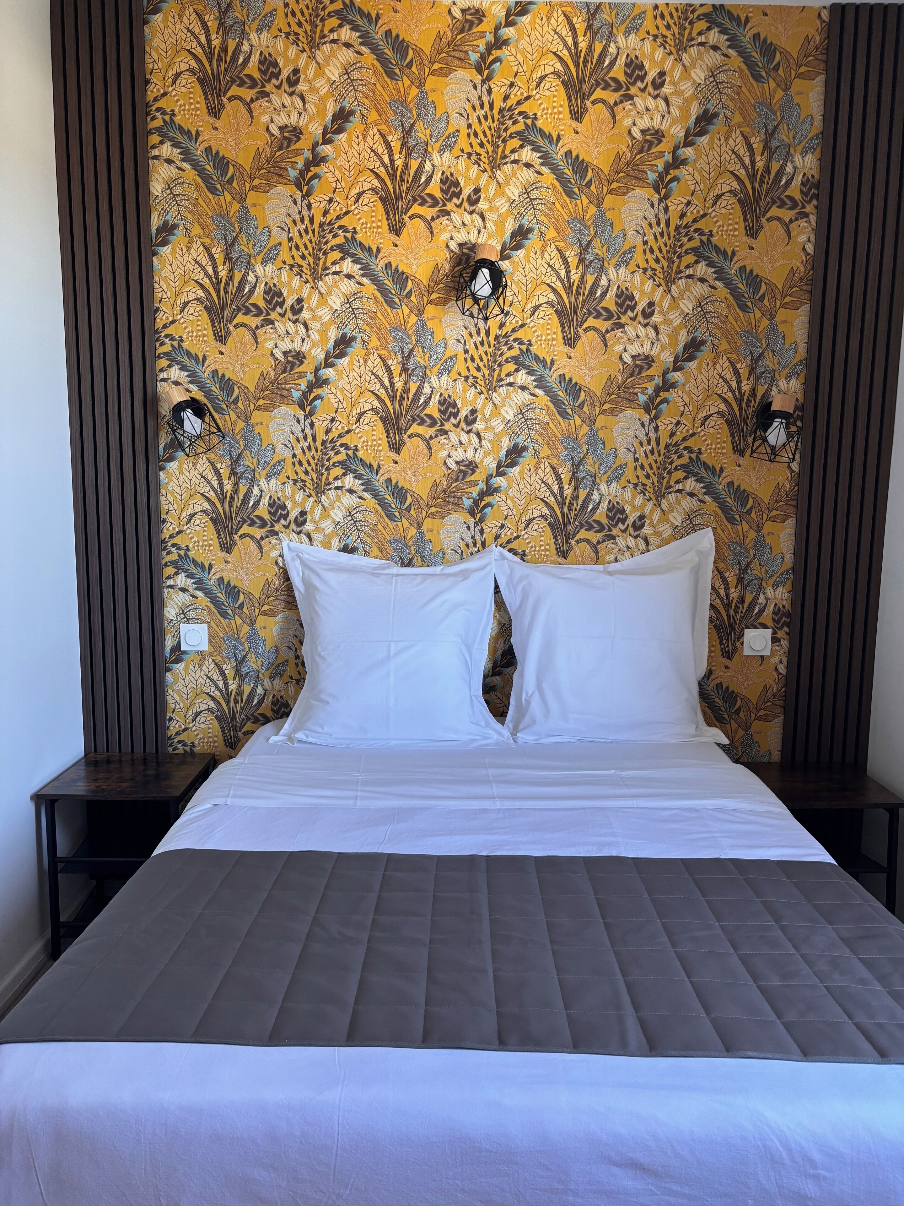 A bed with white linens, a quilted headboard, and a floral-patterned wall behind it.