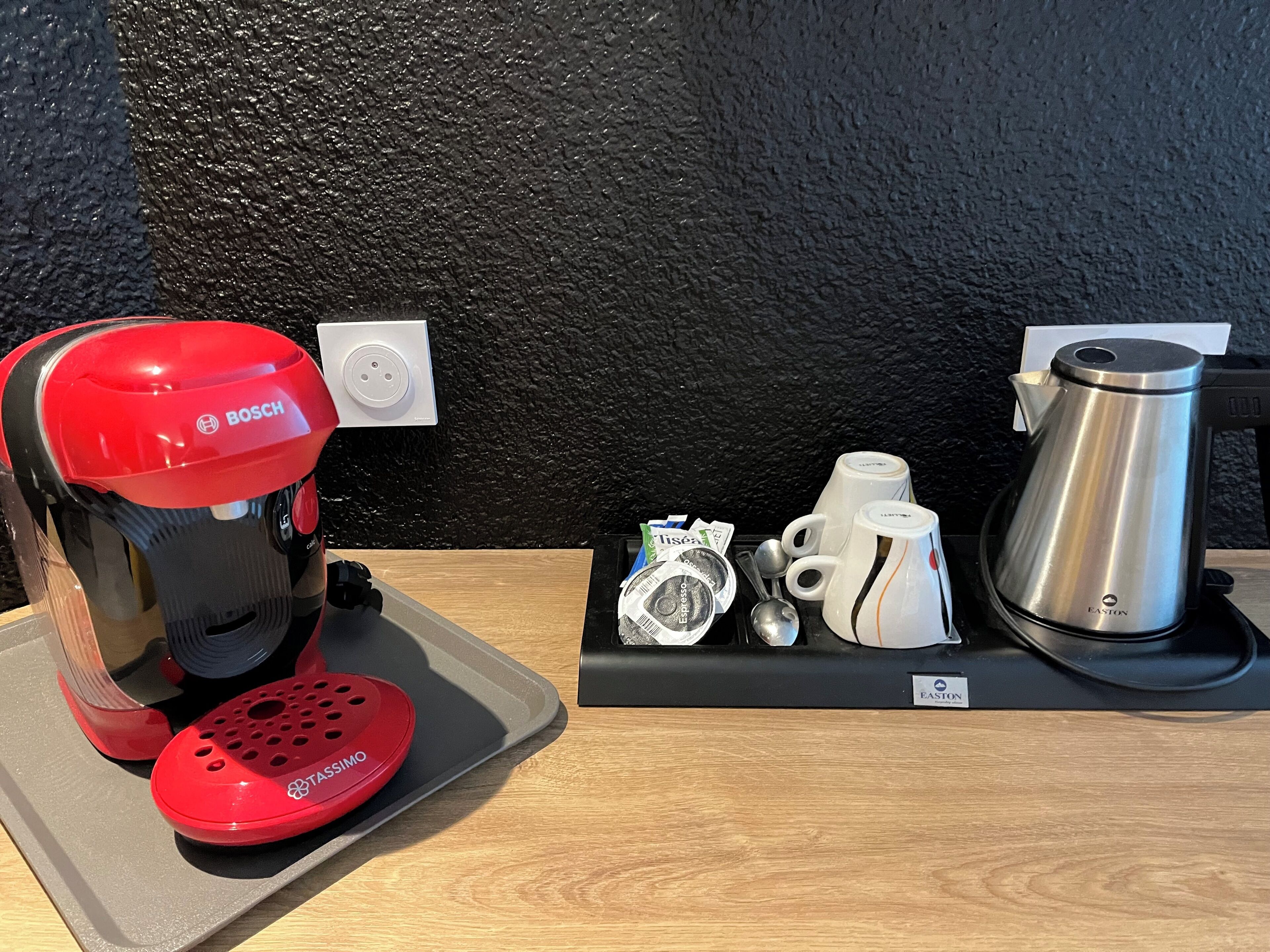 Une machine à café, une bouilloire et une cafetière sont posées sur une table, avec une prise de courant visible à l’arrière-plan.